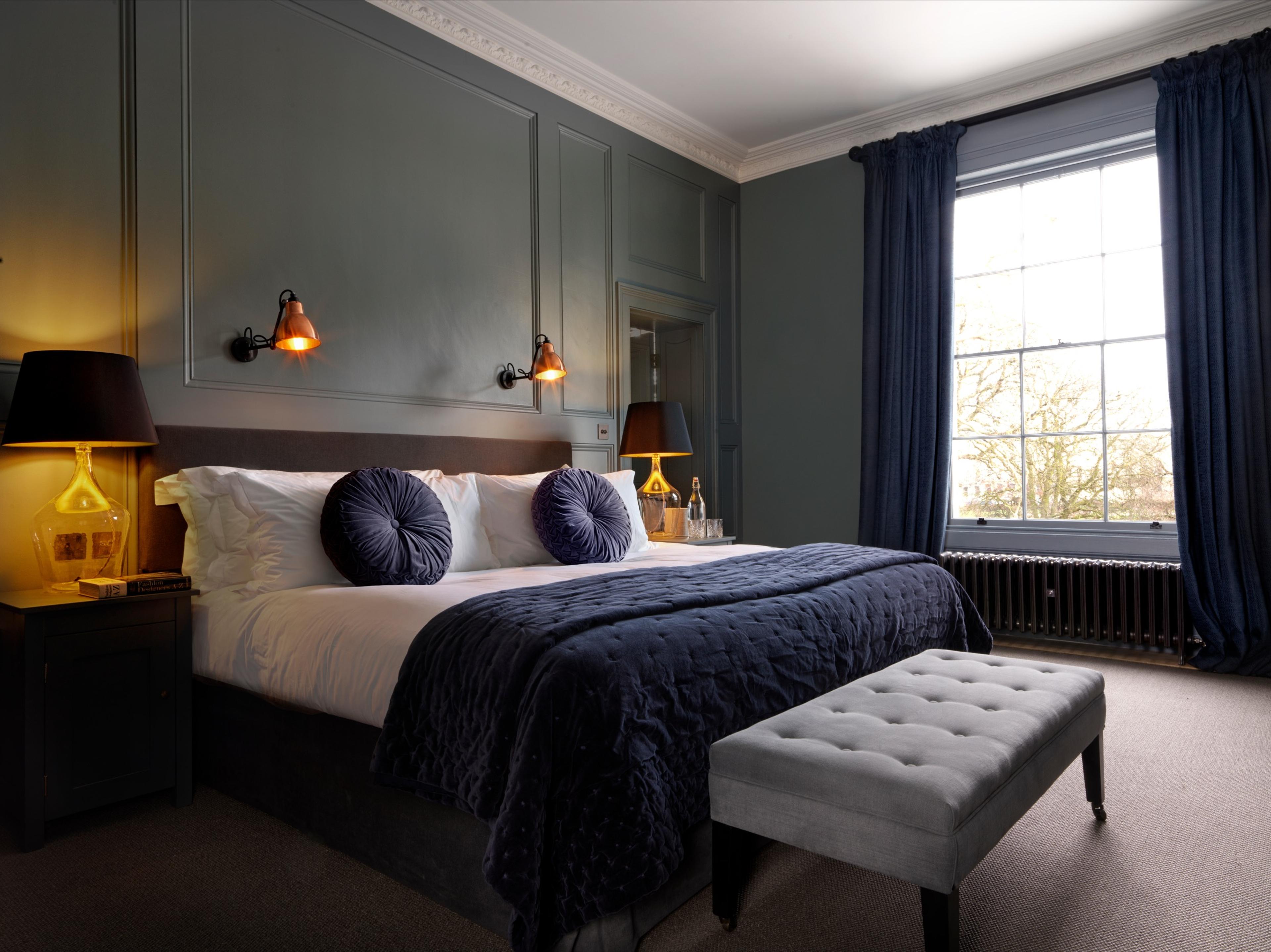 A bedroom with grey-green walls, a navy upholstered bed and a tall sash window looking out onto bare trees.