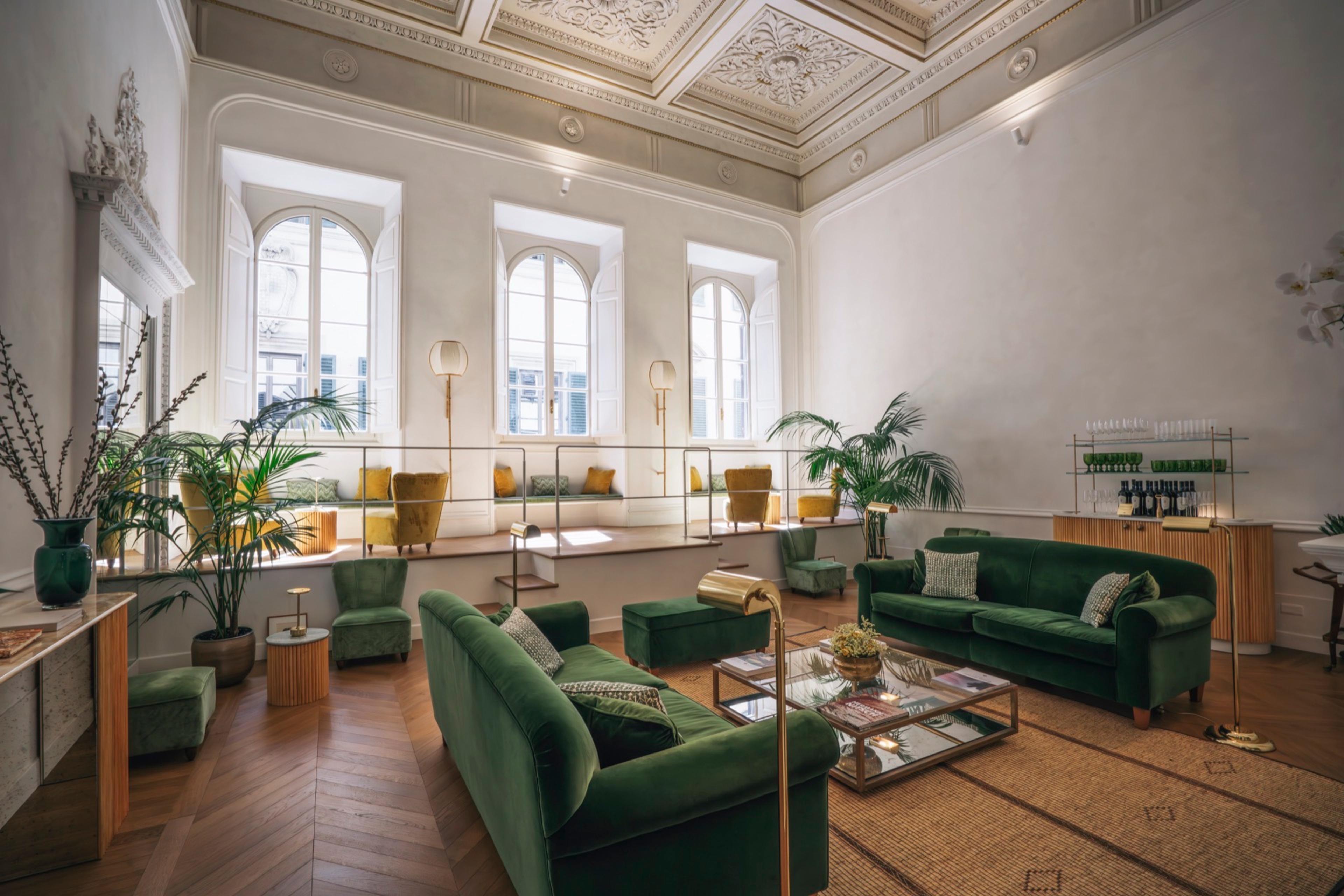 A grand living room with a frescoed ceiling and three tall arched windows, green velvet sofas, potted palms and a built-in bar, seen from a raised step.