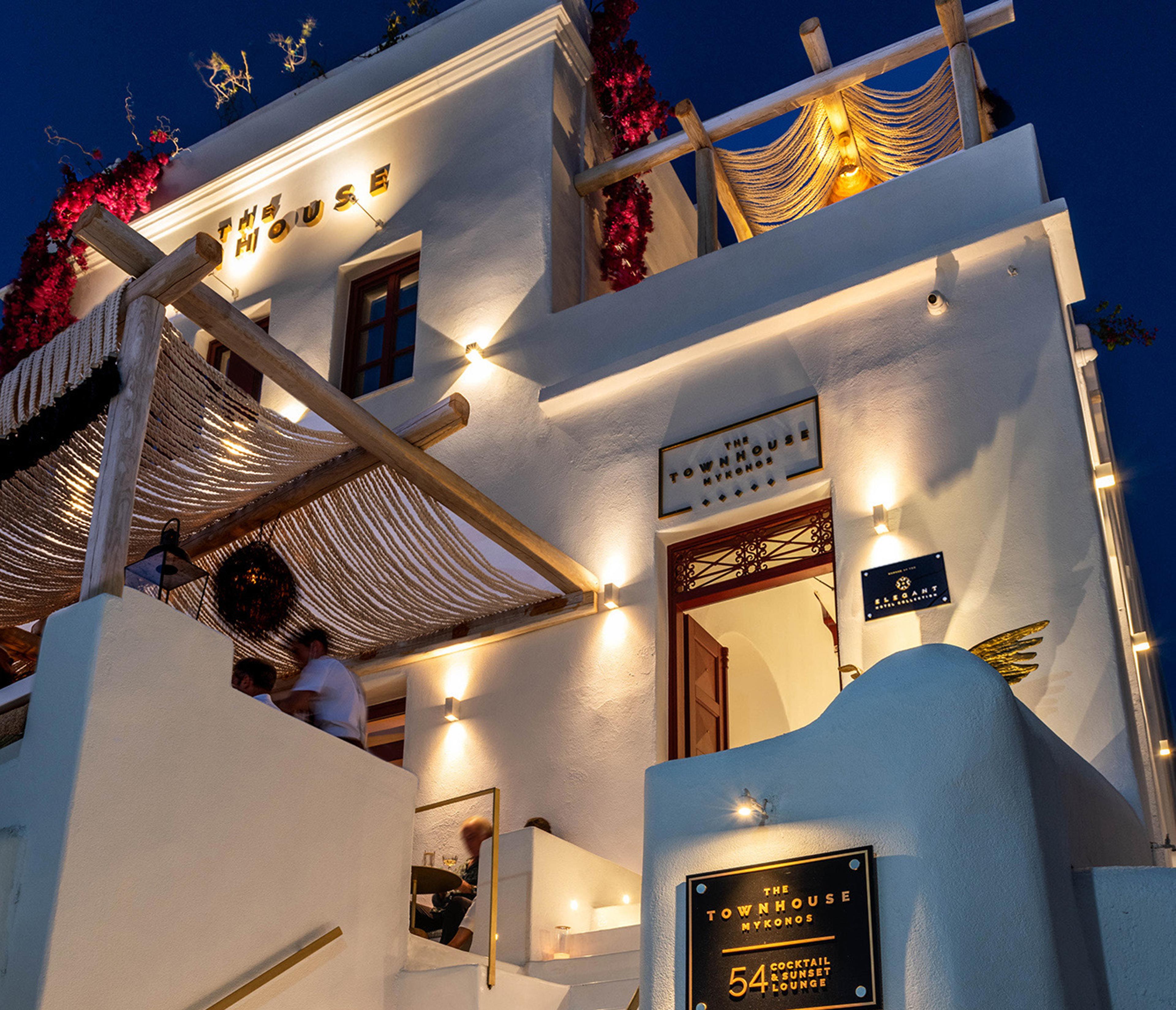 The hotel's white exterior lit up at night, with its sign above an open red door and two people seated on a balcony.