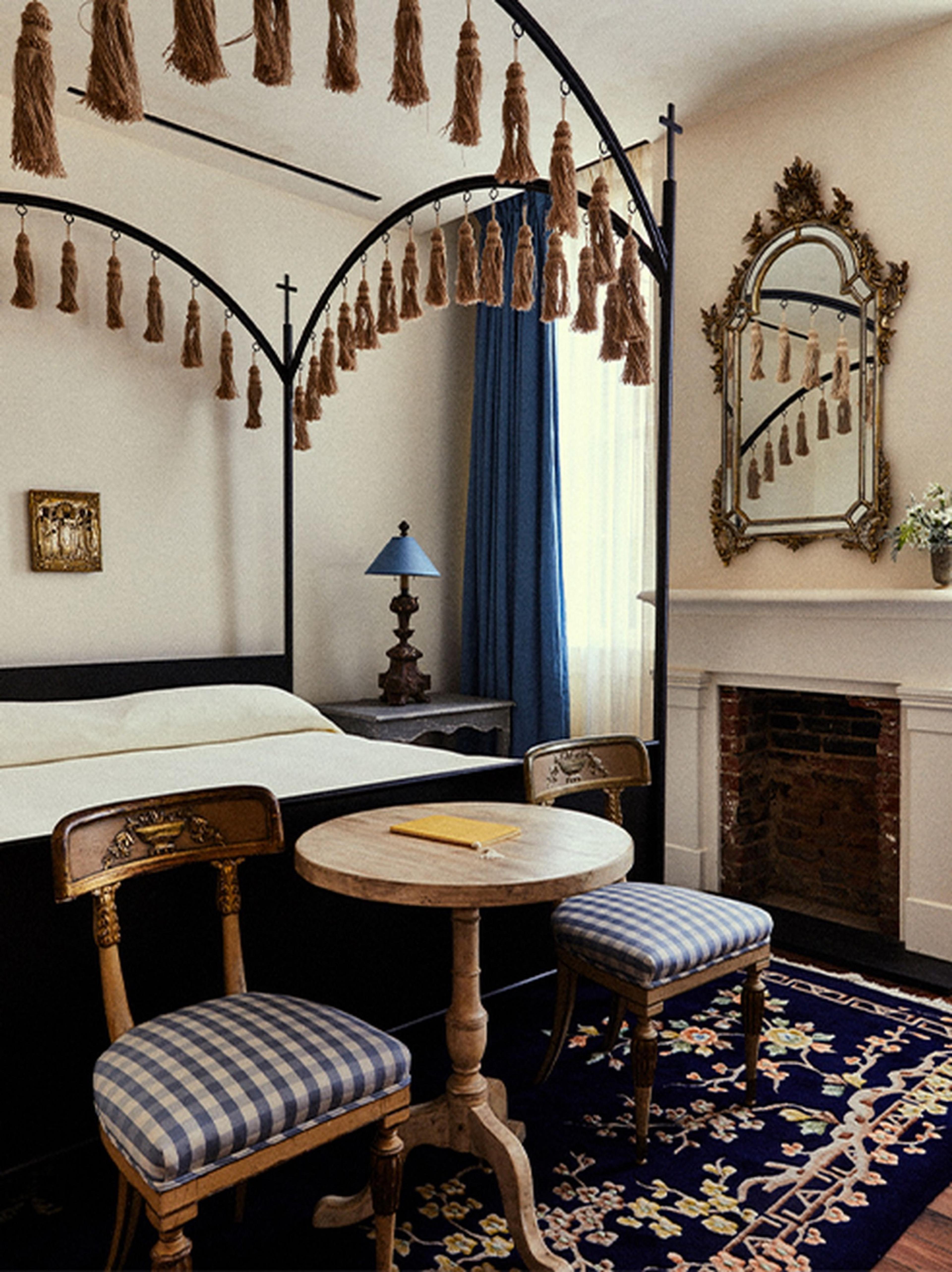 A four-poster bed hung with rope tassels and blue curtains, a round table and two chairs in front of it, and a gilt mirror over a fireplace.