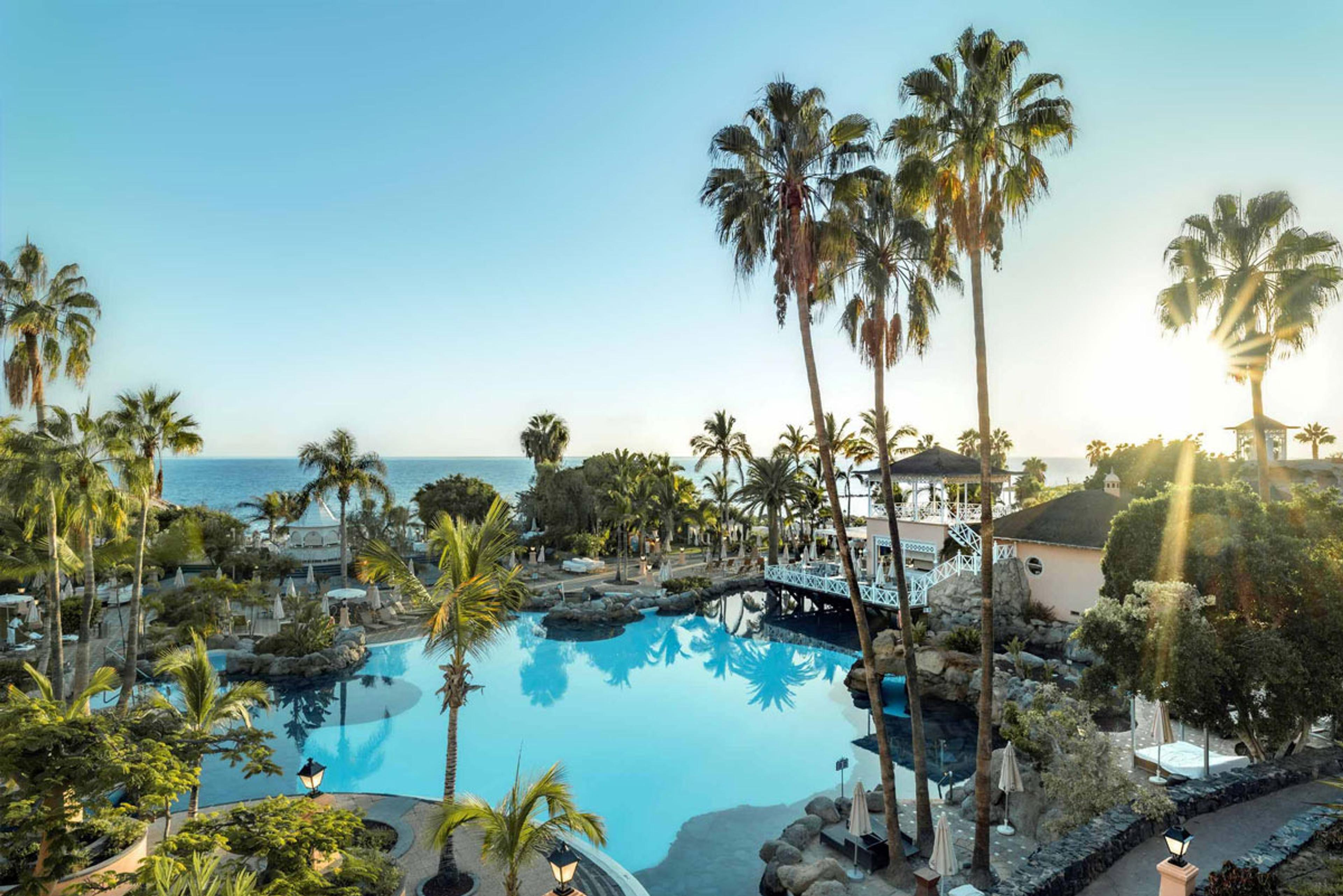 An elevated view over a lagoon-shaped pool ringed by palm trees, with a pergola bridge and the sea beyond at sunset.