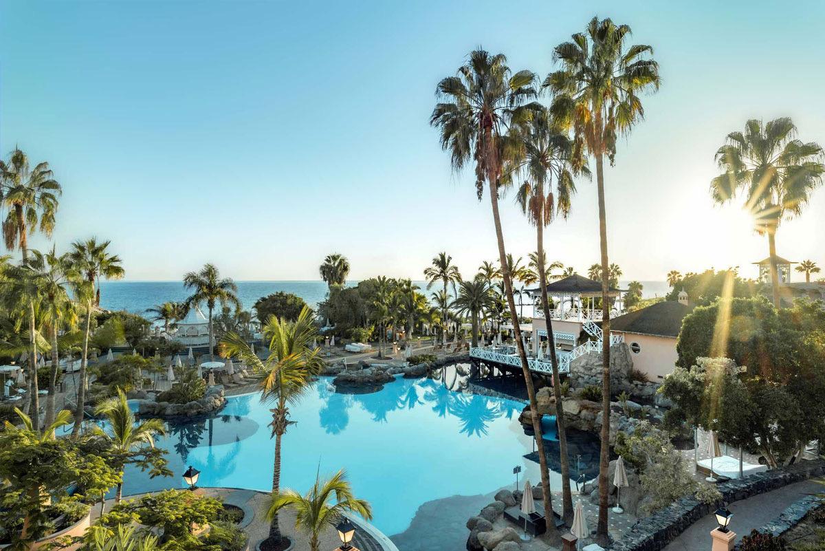 An elevated view over a lagoon-shaped pool ringed by palm trees, with a pergola bridge and the sea beyond at sunset.