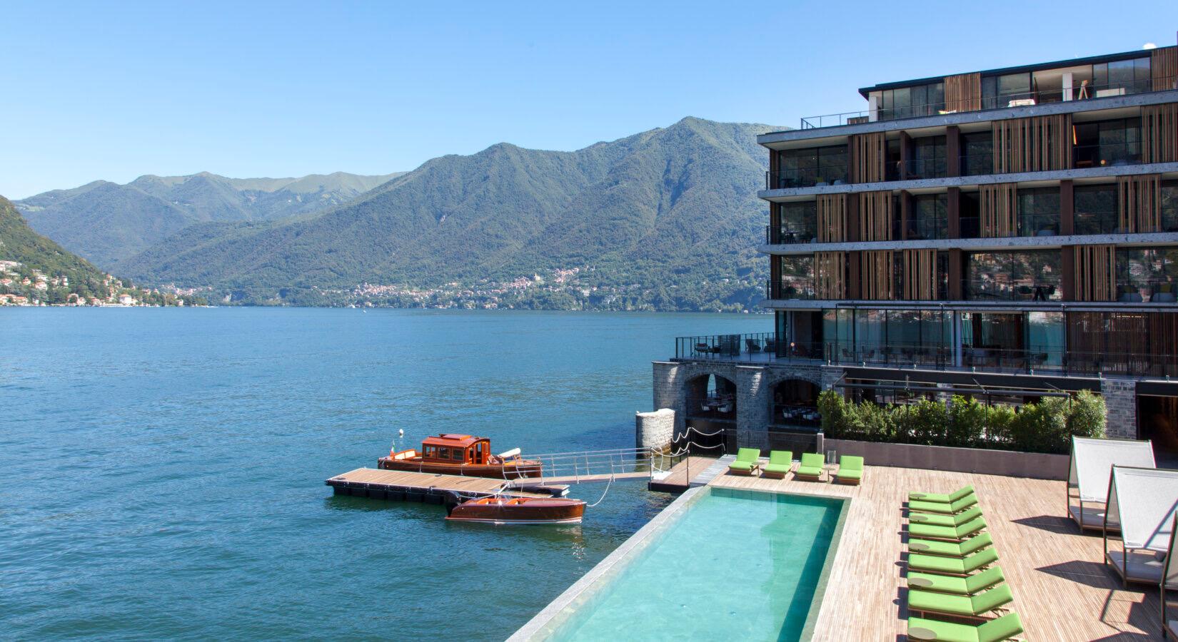 A hotel building beside a lake, seen from the water, with an infinity pool, sun loungers and two wooden boats moored at a jetty, mountains rising behind.