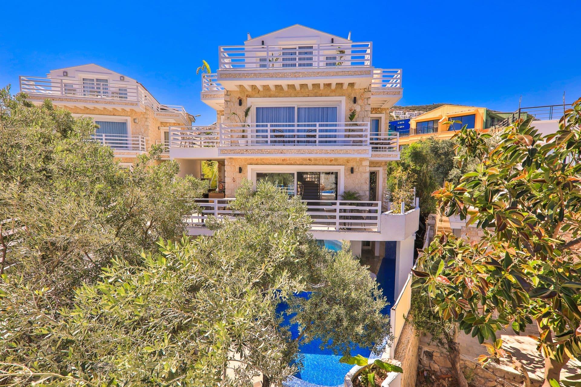 A stone-and-white hotel building rises several storeys above two swimming pools, framed by olive trees under a blue sky.