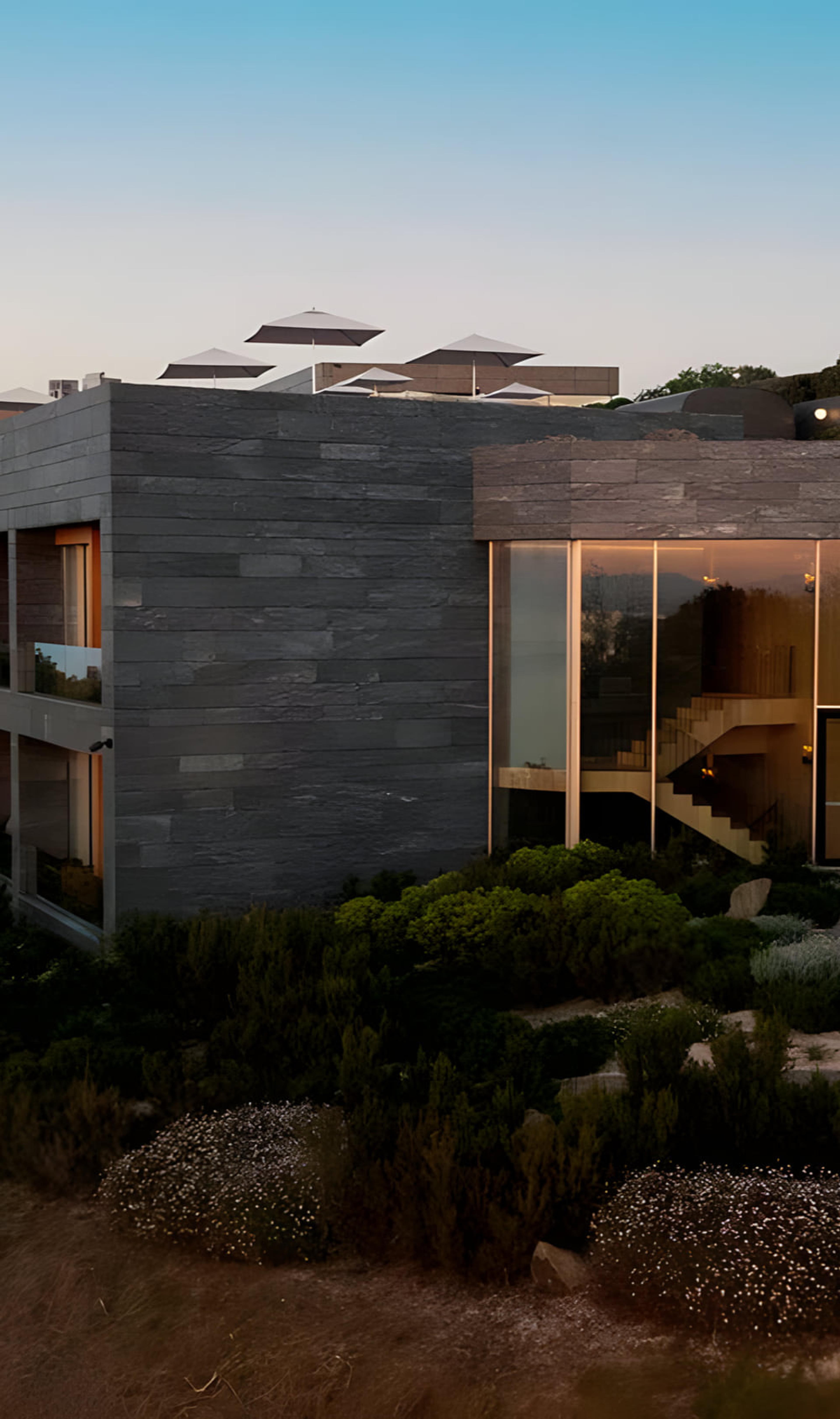 The corner of a stone-clad hotel wing at dusk, its glass end wall showing an interior staircase, white umbrellas on the roof terrace above.