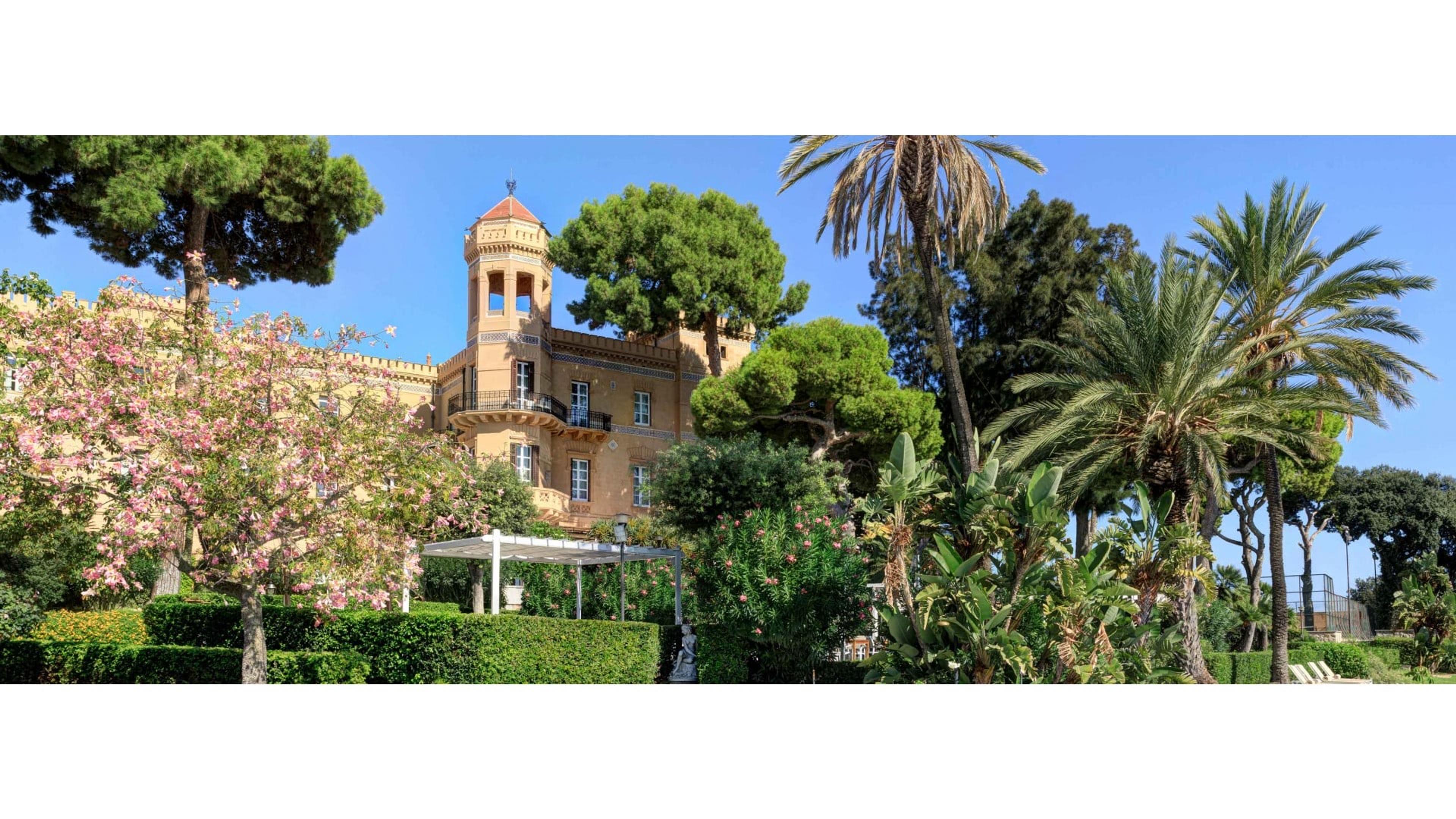 A turreted ochre building rises behind a garden of pine and palm trees, with a blossoming pink tree and clipped hedges in the foreground.