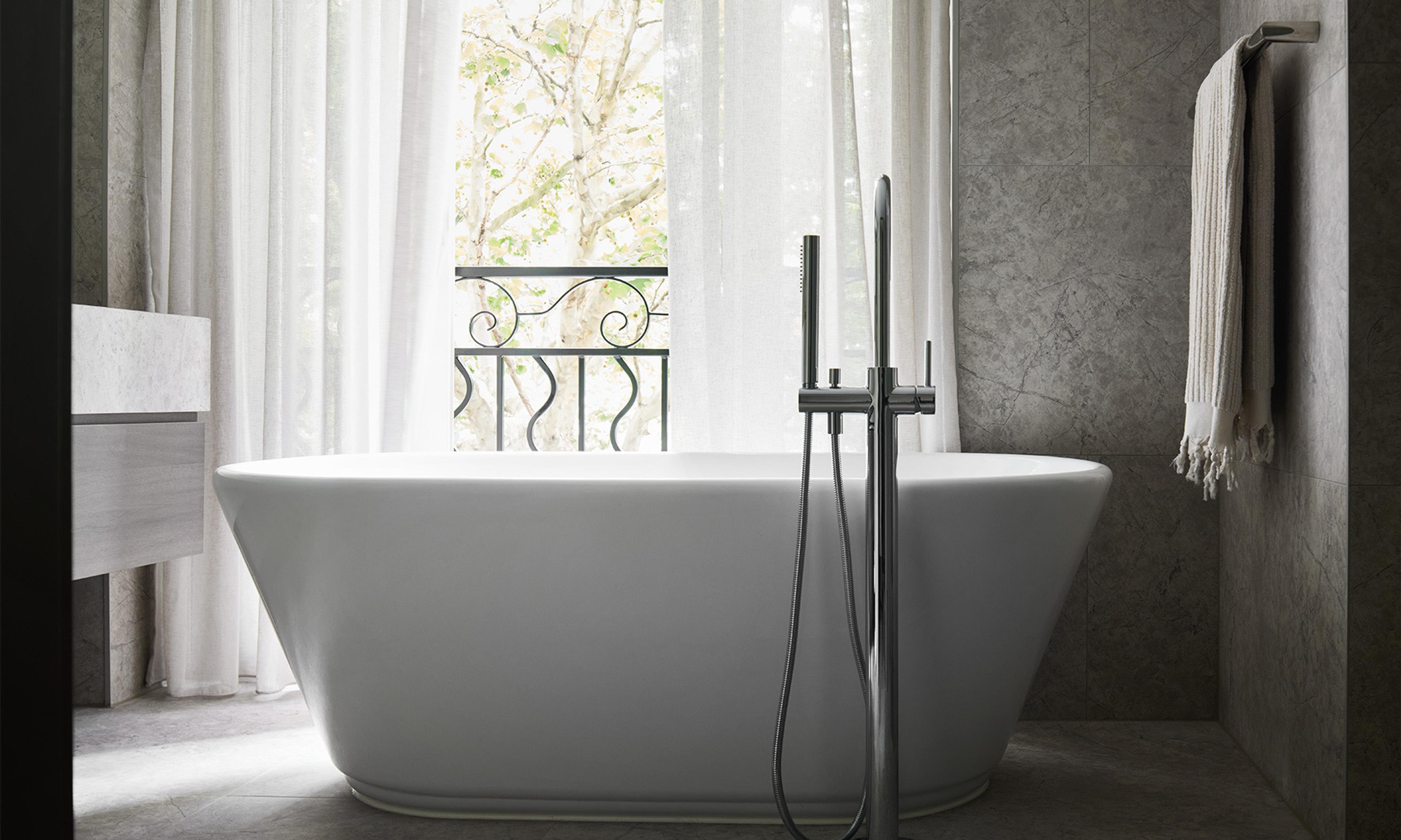 A freestanding bathtub facing a window with sheer curtains open to a wrought iron balcony and trees outside.