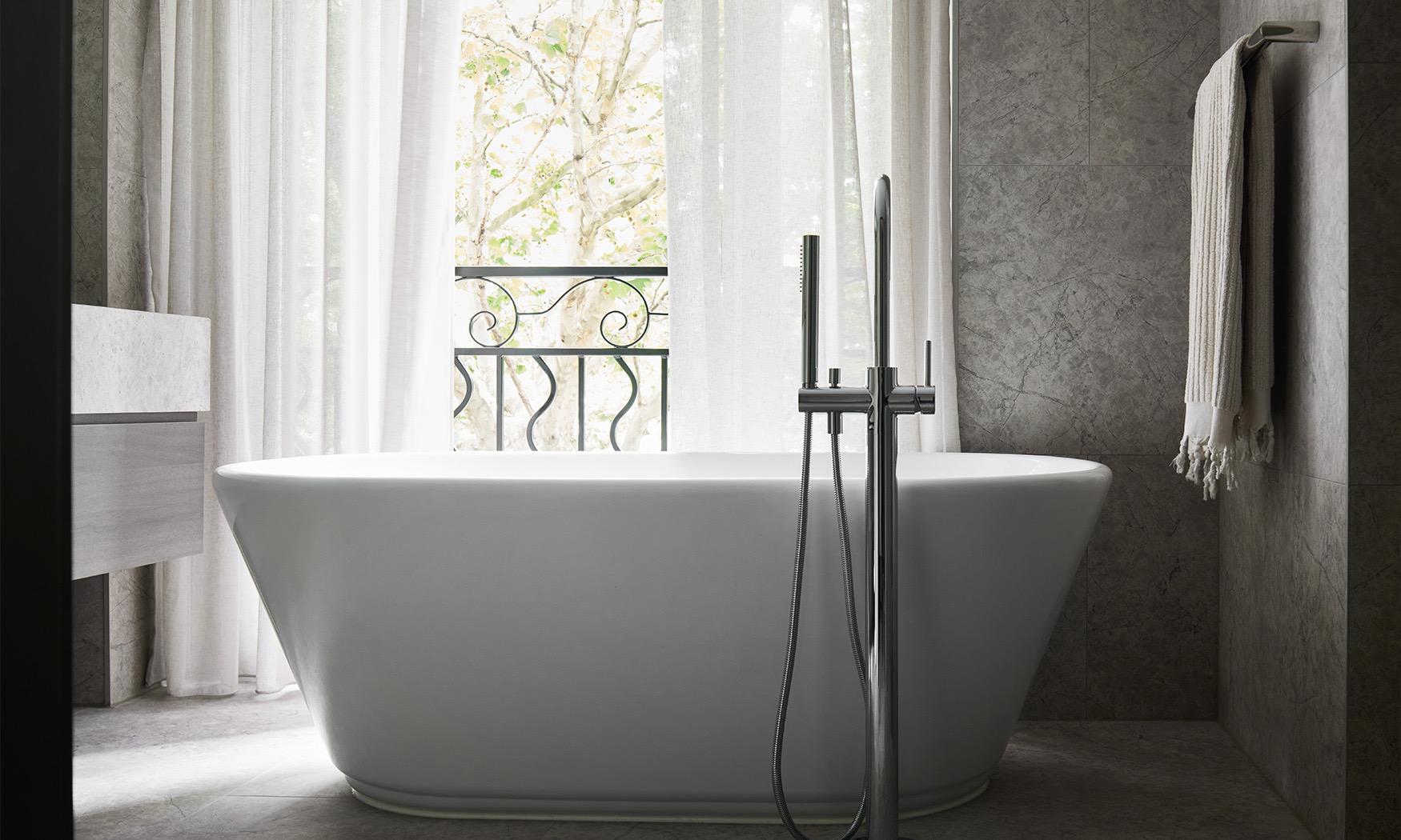 A freestanding bathtub facing a window with sheer curtains open to a wrought iron balcony and trees outside.