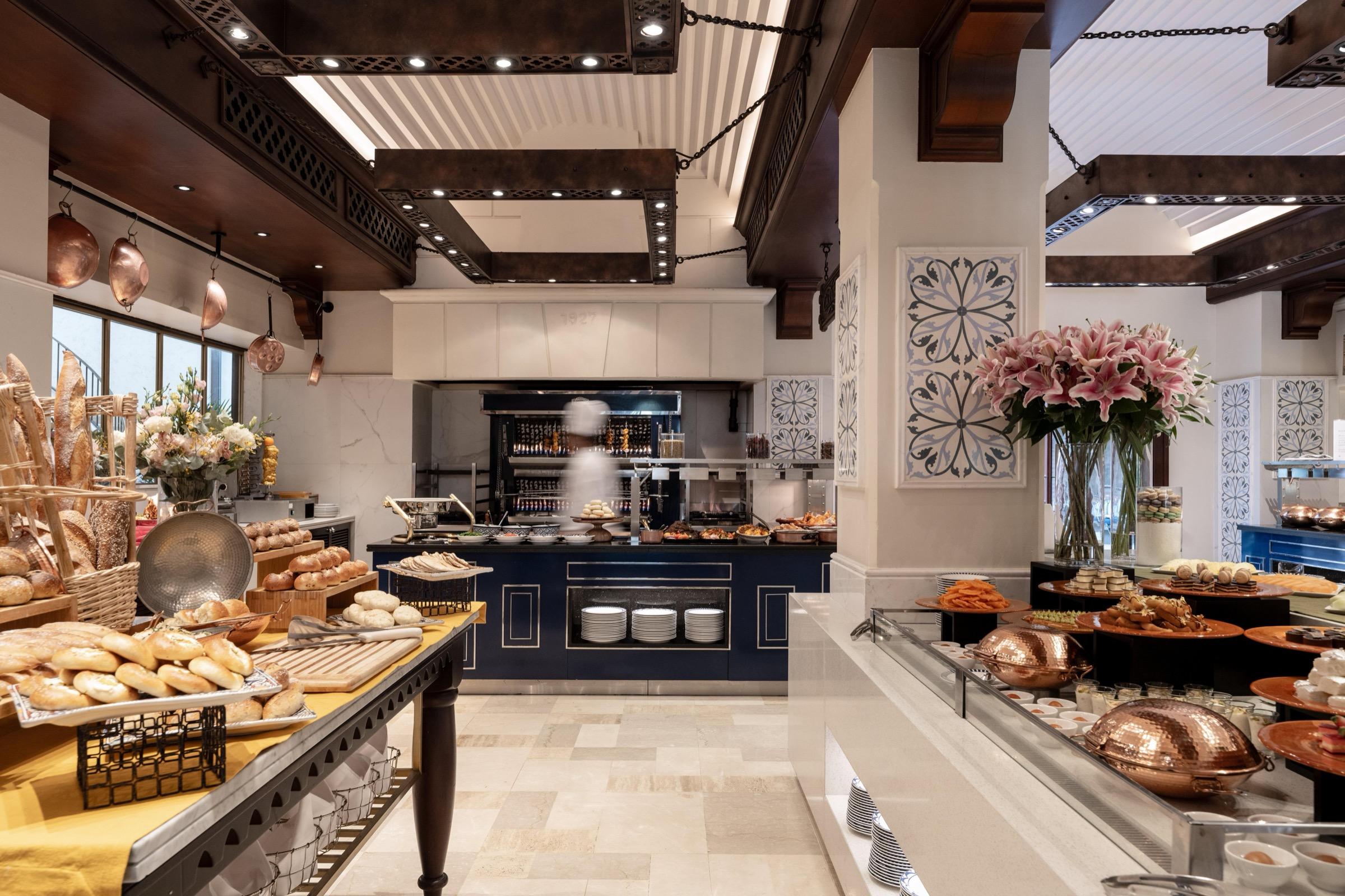 A hotel breakfast buffet with baskets of bread and pastries, copper serving domes, and a large vase of pink lilies.