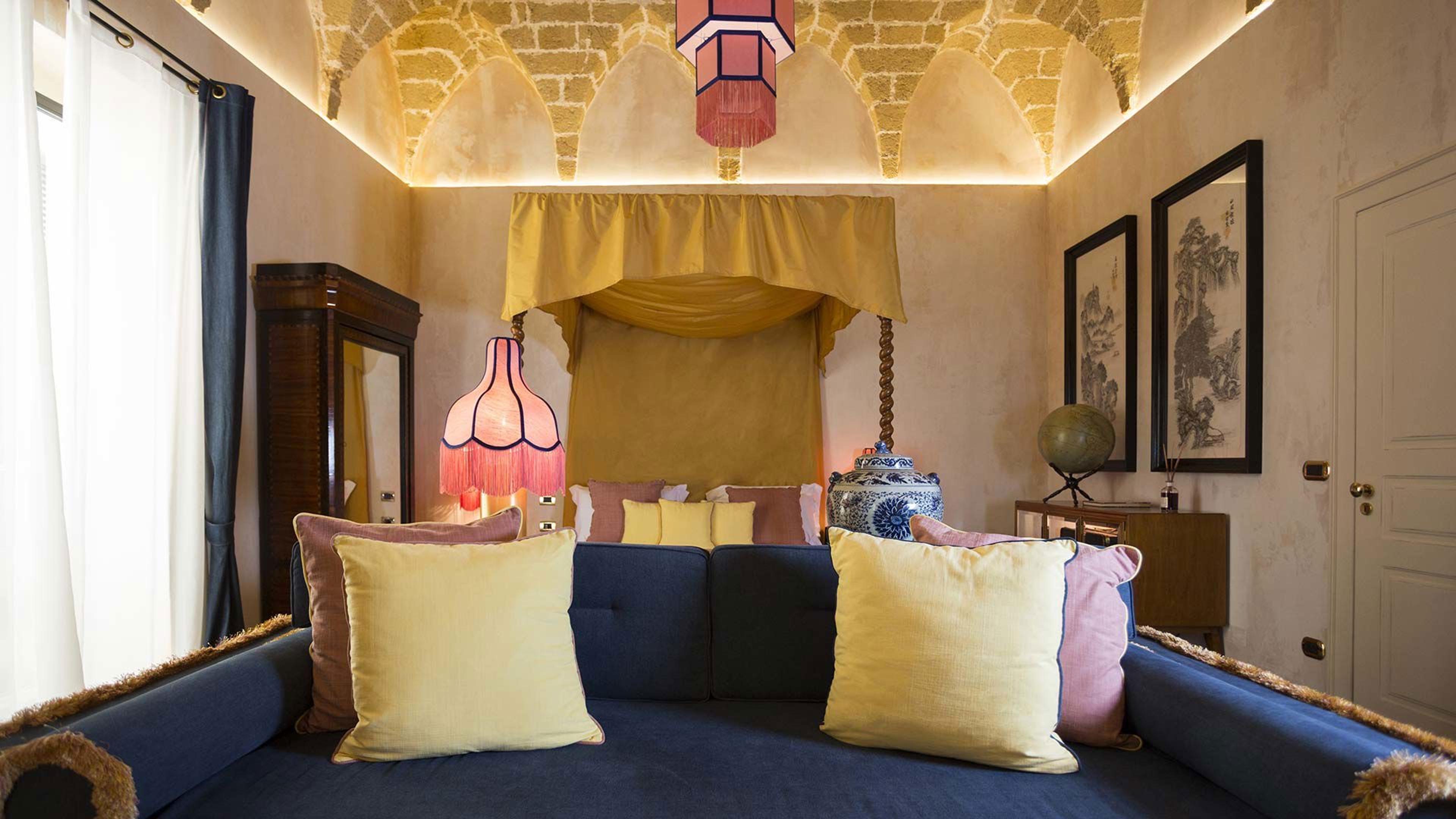 A bedroom with a vaulted stone ceiling, a hanging paper lantern, a four poster bed with a mustard canopy, a blue upholstered daybed with patterned cushions, and a blue and white ginger jar on a cabinet.