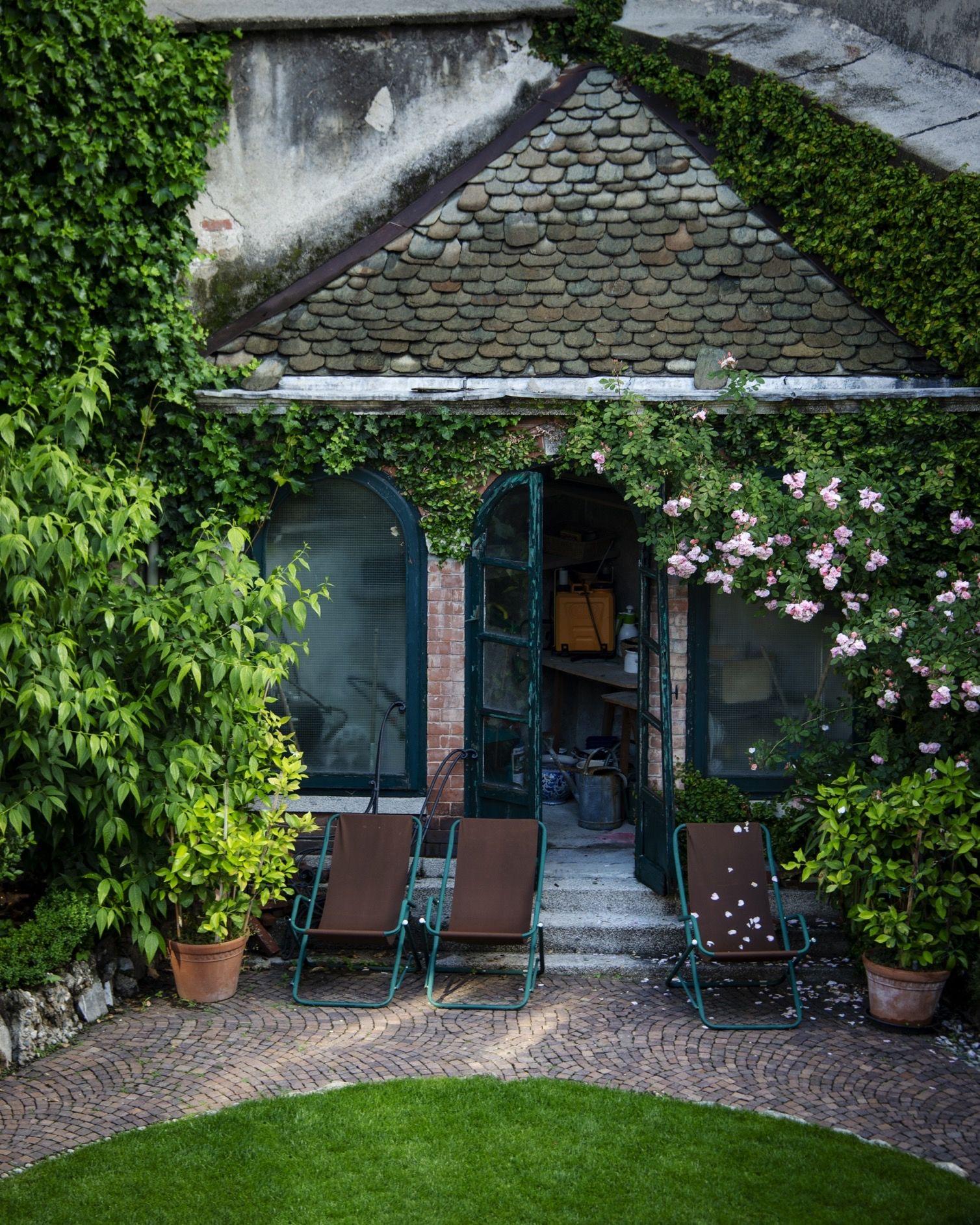 deck chairs and potted plants on a brick terrace outside an ivy-covered stone outbuilding with a tiled roof and climbing roses