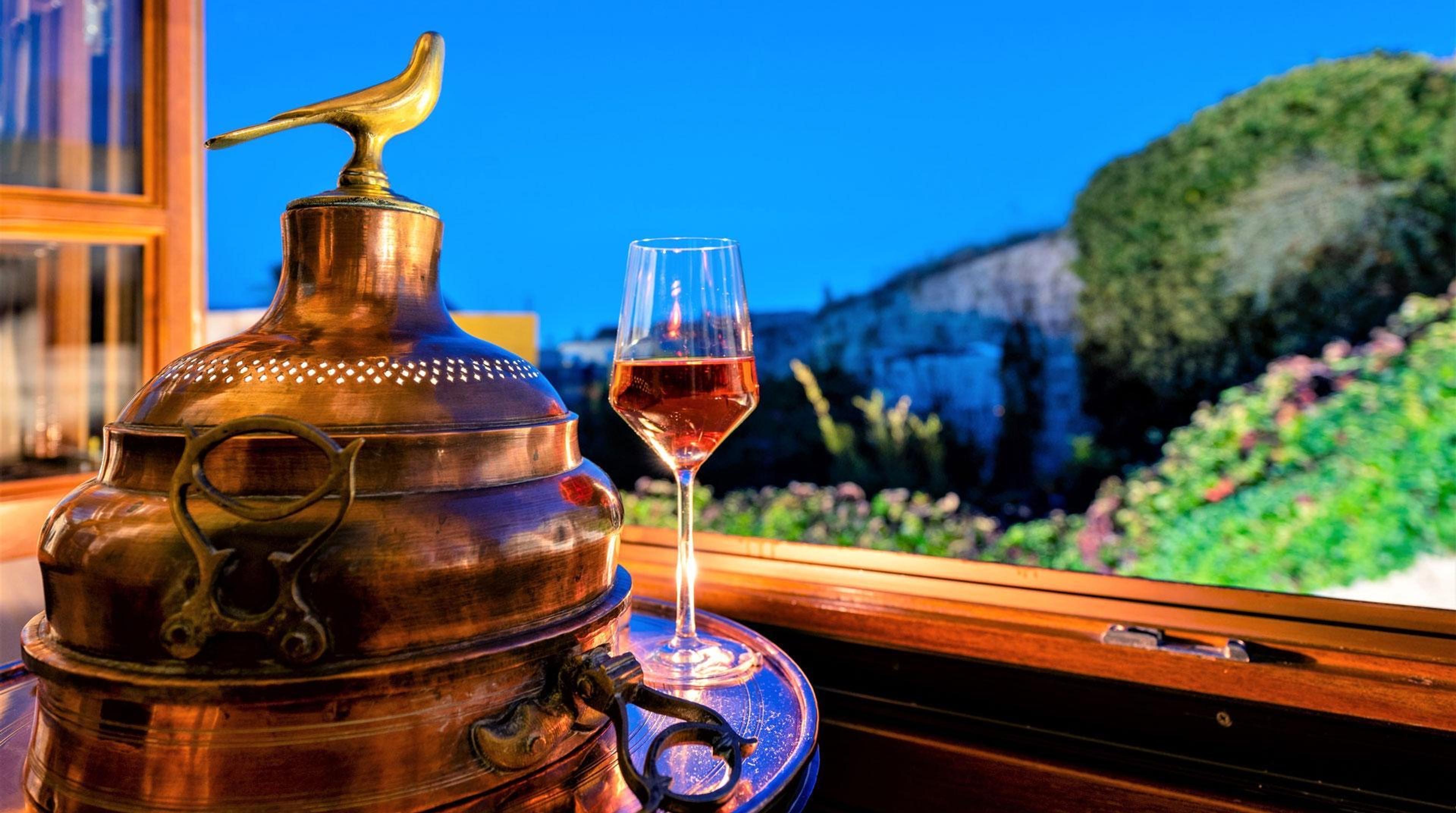 A copper still and a glass of rose wine on a windowsill, with greenery and a stone wall beyond.