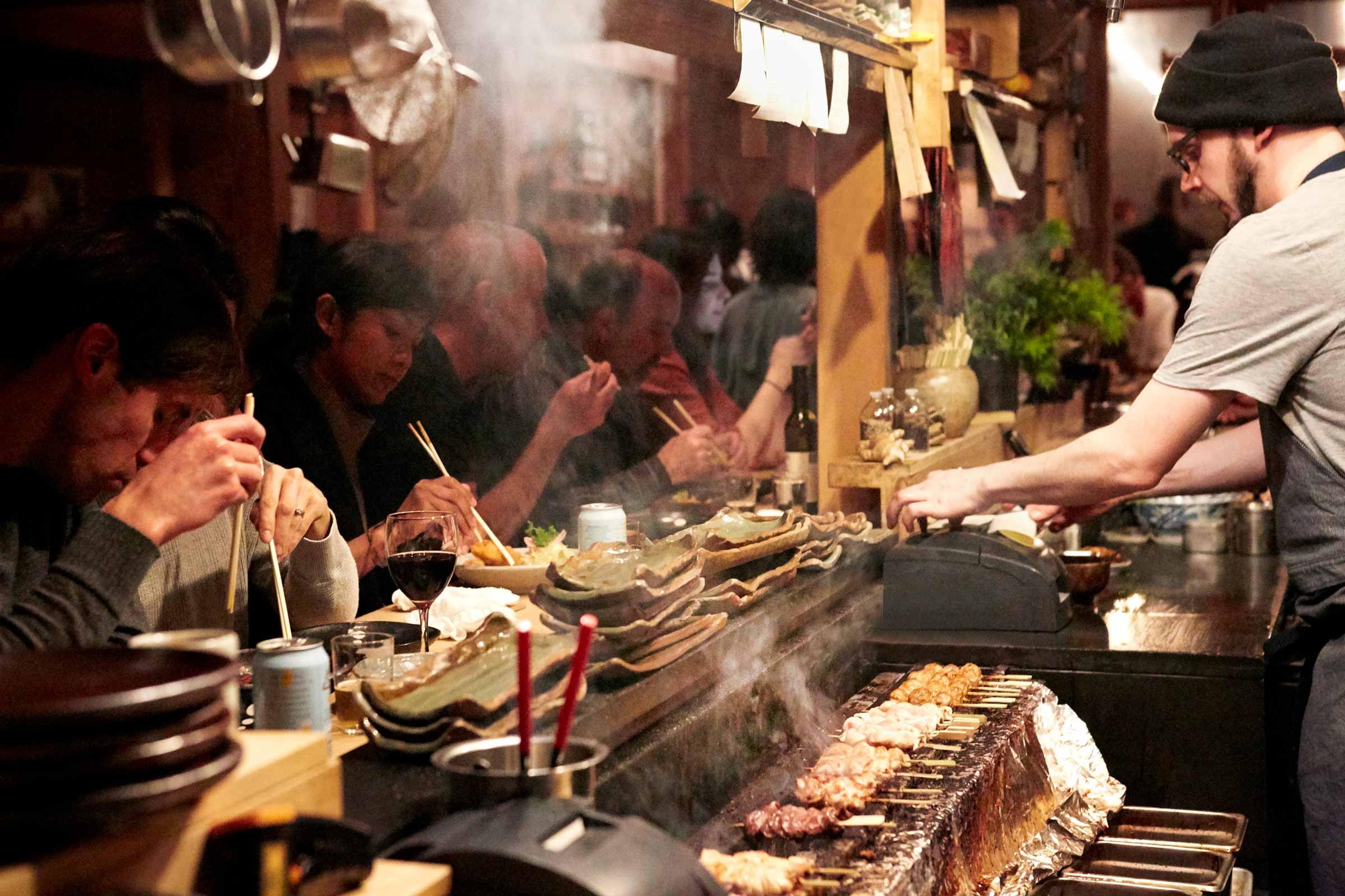 Diners at a wooden counter in front of a smoking charcoal grill, cooks turning skewers behind it