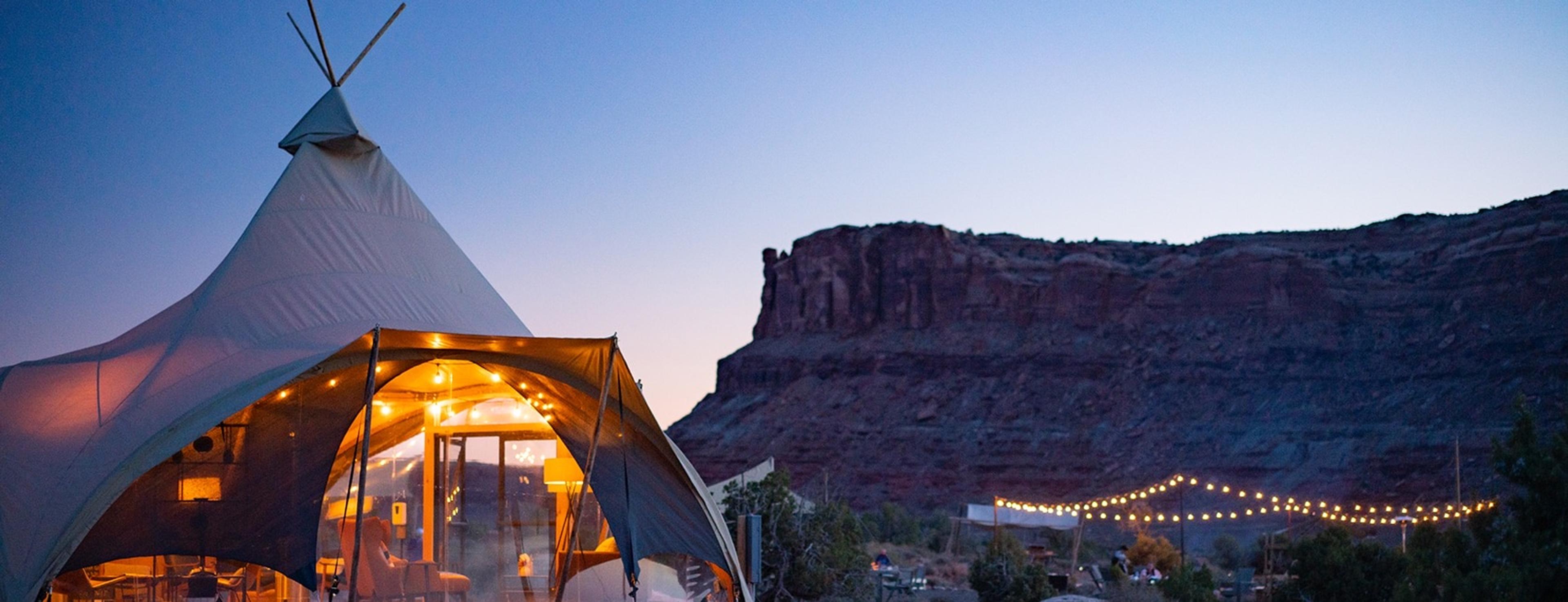 A canvas tipi at dusk, its sides glowing from the lamps inside, with a red sandstone butte rising behind and a line of string lights along the ground.