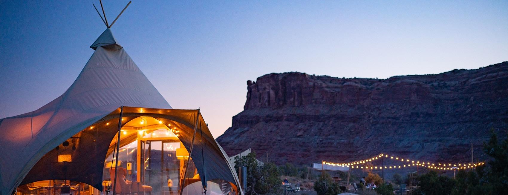 A canvas tipi at dusk, its sides glowing from the lamps inside, with a red sandstone butte rising behind and a line of string lights along the ground.