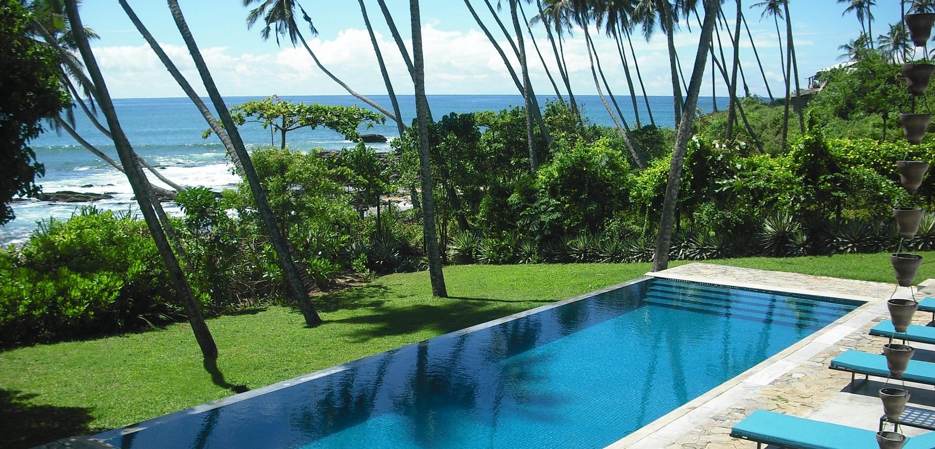 A turquoise infinity pool lined with palm trunks overlooks a rocky cove and open sea, with blue sun loungers along one edge.