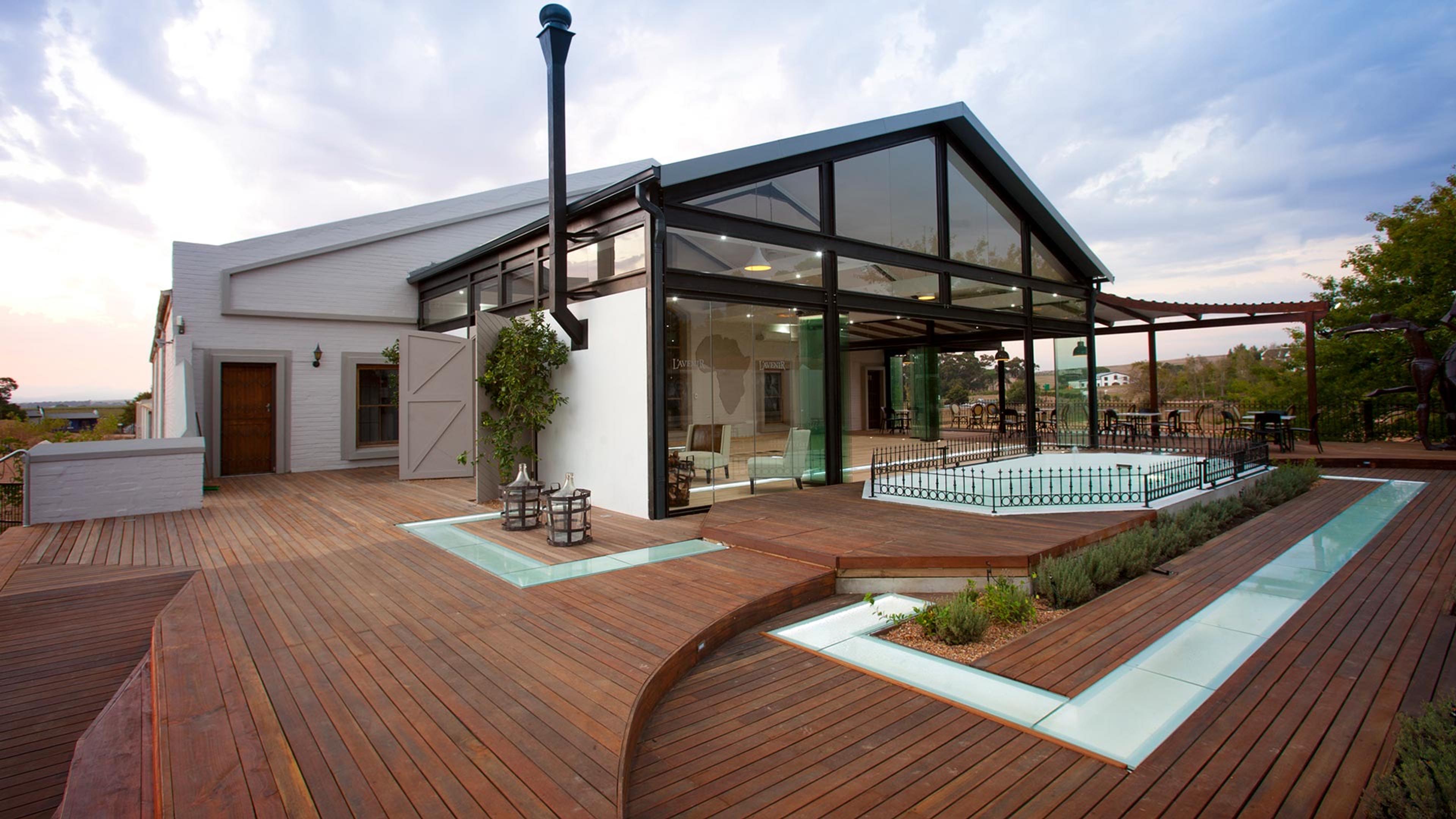 A white lodge with a black-framed glass gable, a tall flue, and a curved timber deck in front, with a pool behind a railing and a dusky sky with cloud.