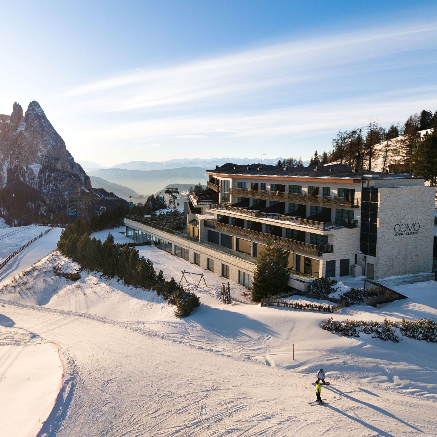 A hotel with a stone and timber facade stands on a snowy slope beneath a rocky peak, with two snowboarders riding past in the foreground.