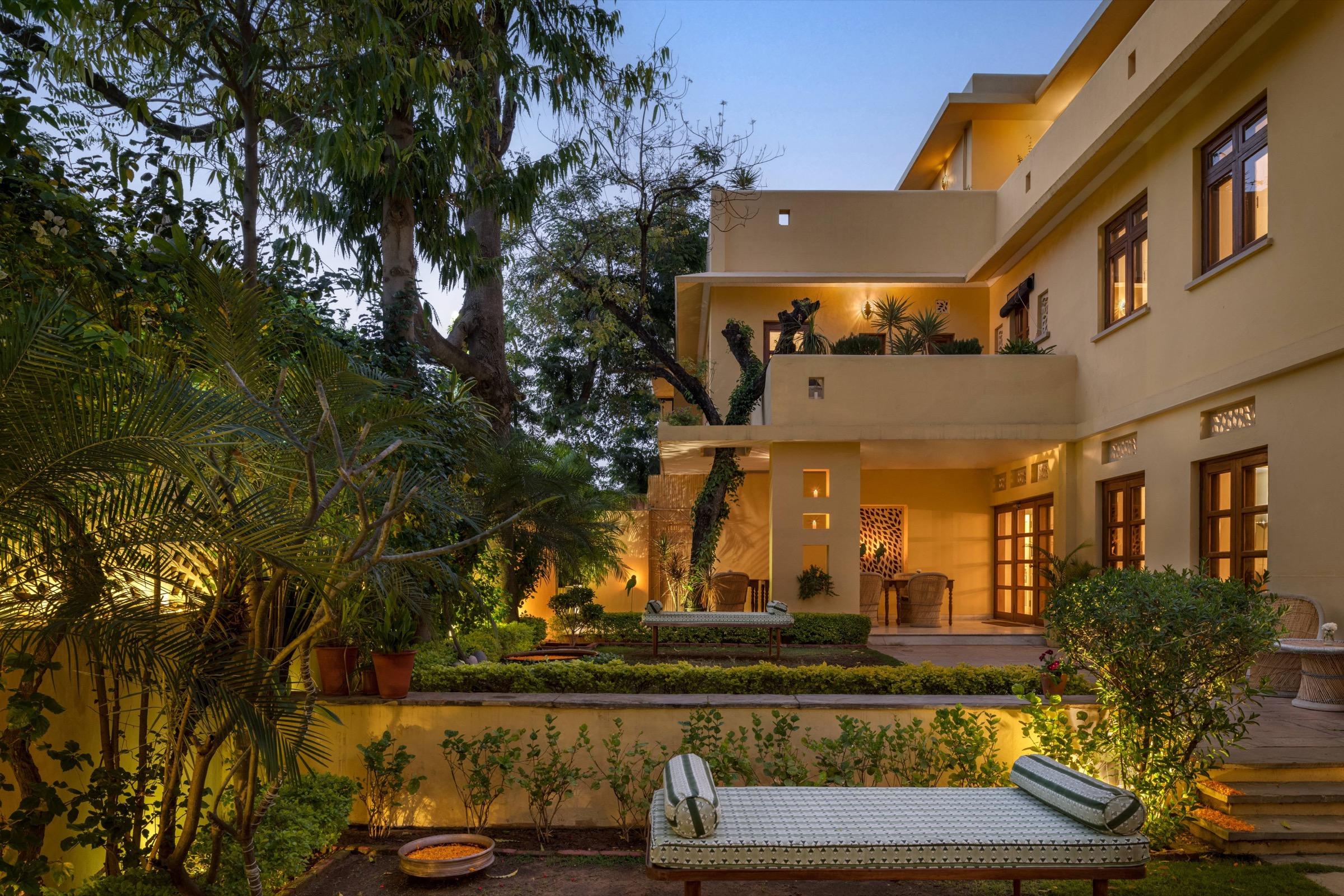 A yellow building lit up at dusk with a daybed and garden in the foreground, steps leading up to a terrace and trees framing the view.