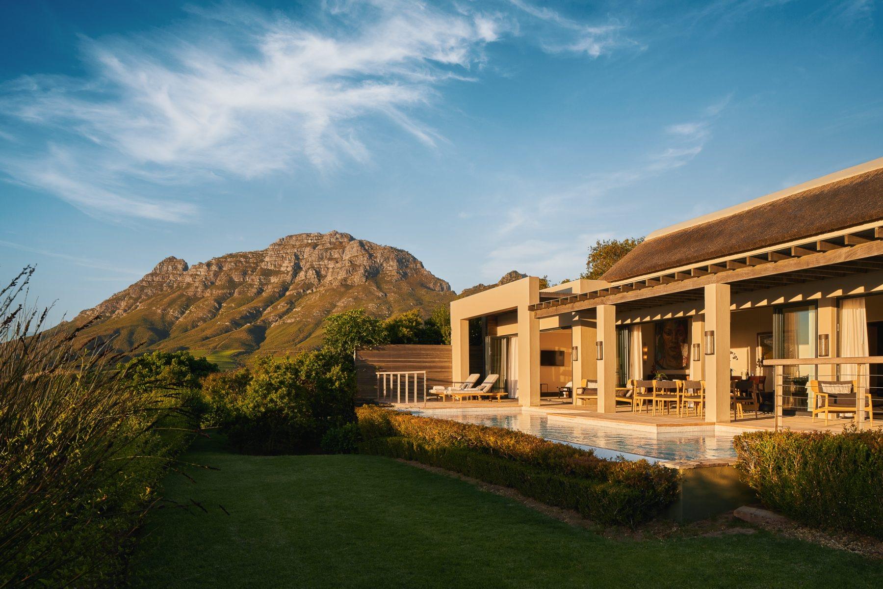 A thatched lodge with a long pool and a lawn in front, below a rocky mountain in evening sun.