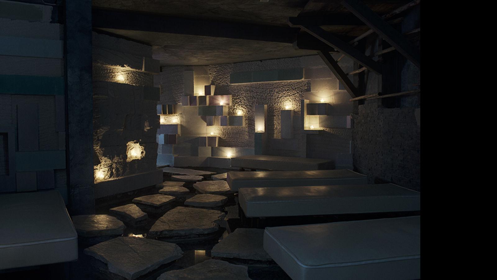 A candlelit stone lounge with low upholstered daybeds beside a shallow water channel and stepping stones.