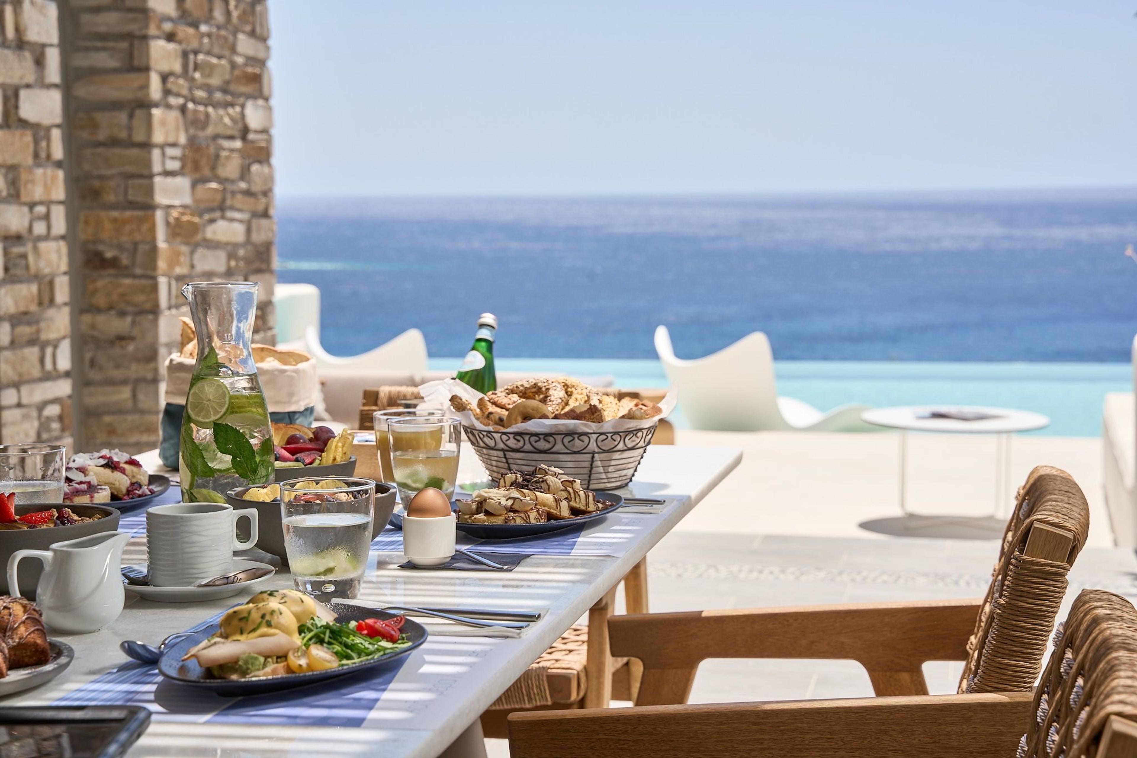 A long white table on a terrace laid for breakfast with bread, fruit, eggs, coffee and juice, with a pool and the sea behind.