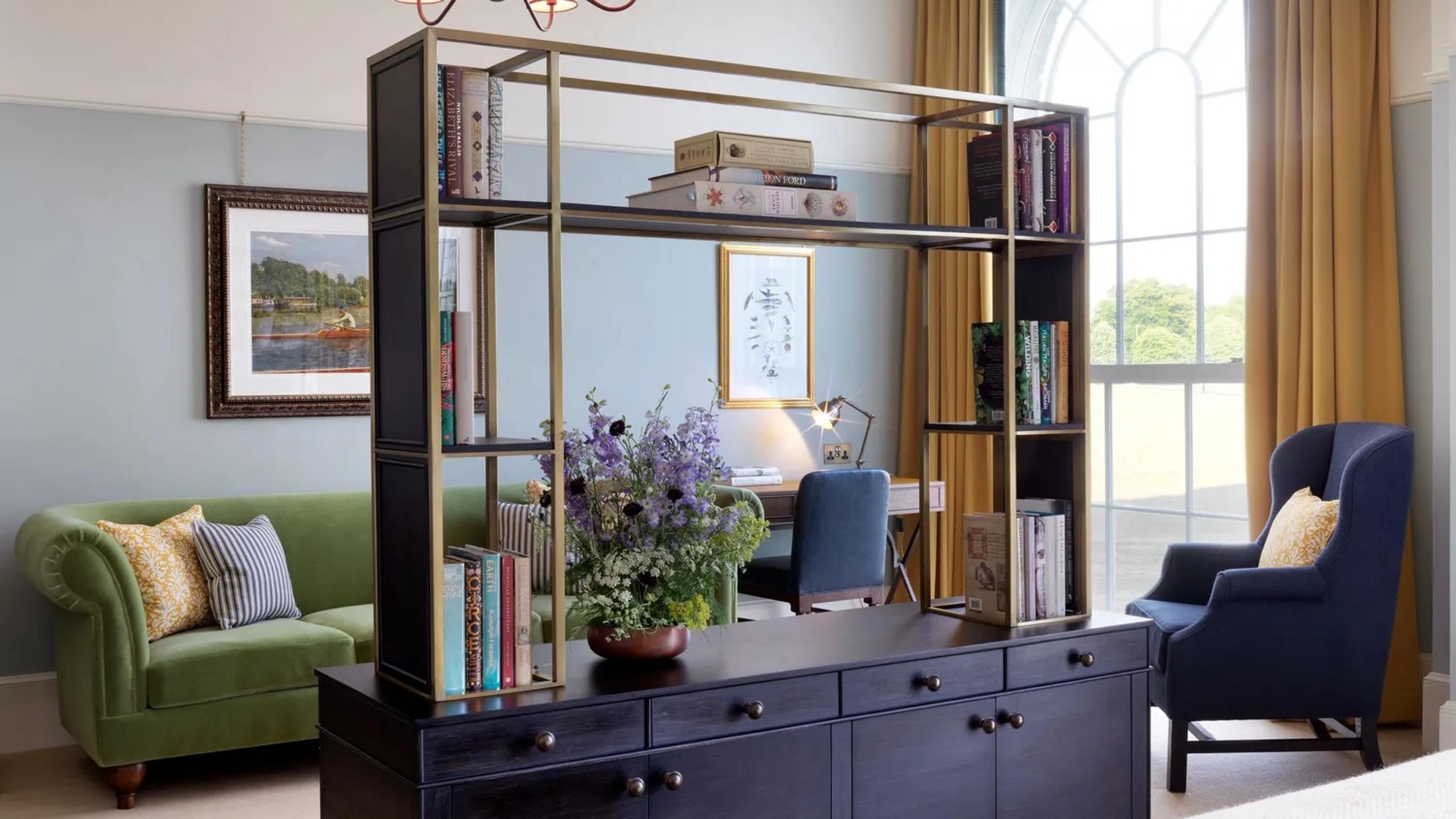 a suite sitting area with an open metal shelving unit of books, a green sofa with patterned cushions, a writing desk with a blue chair, and a tall arched window with mustard curtains