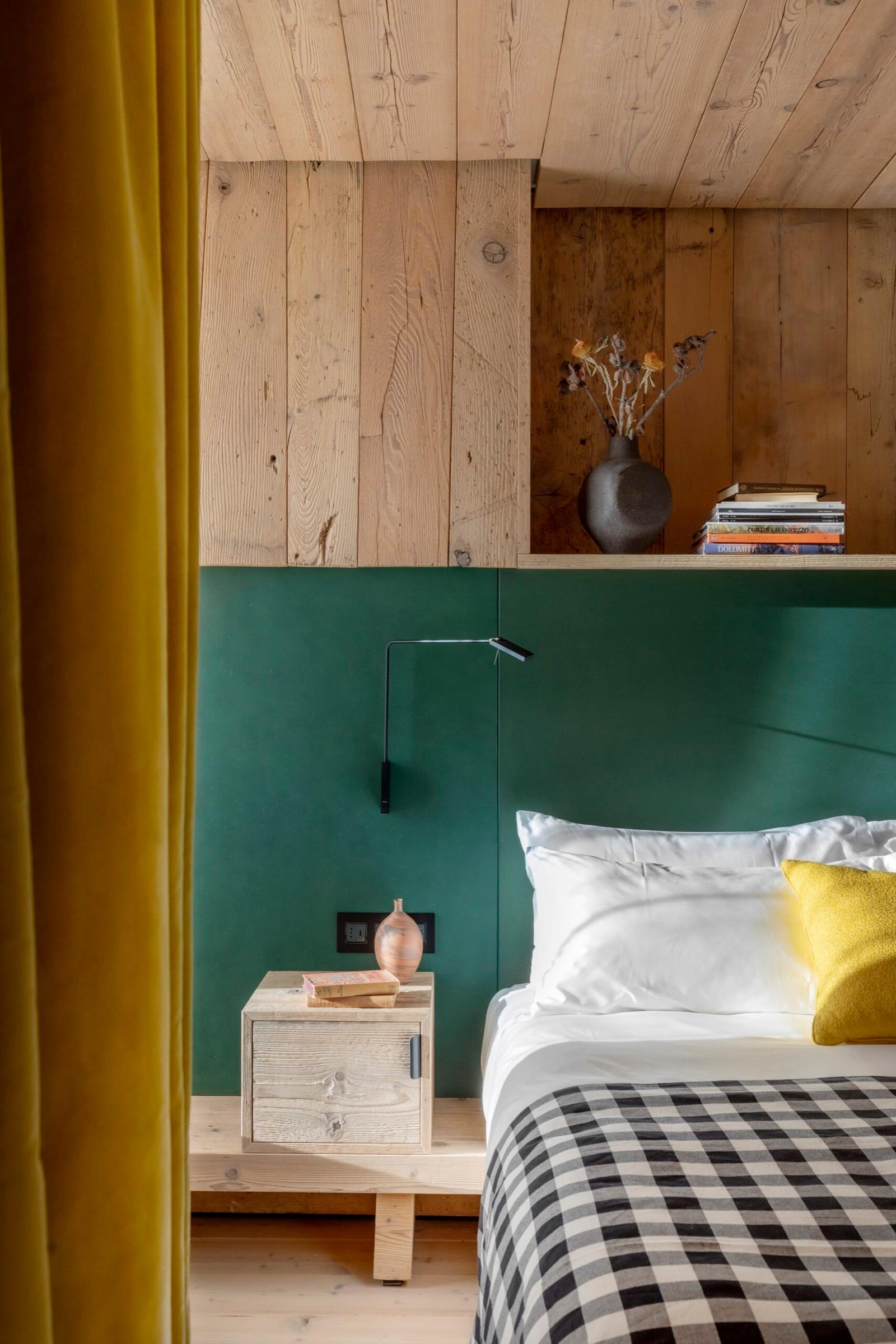A bed with a checked throw and a yellow cushion sits against a dark green headboard, with a vase and books on a shelf above.