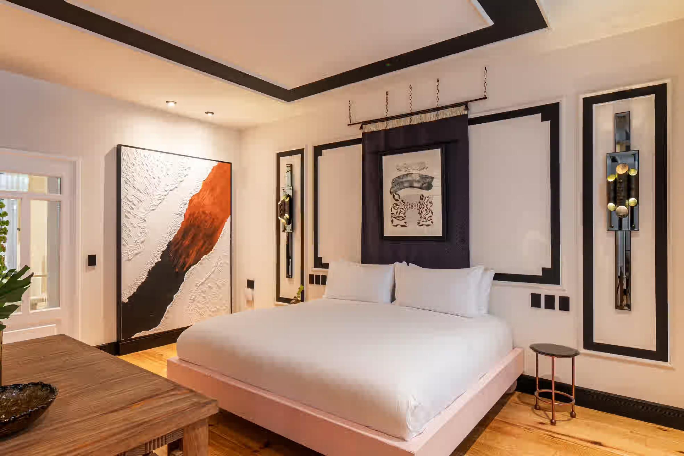 A pink-toned platform bed sits below a large textured canvas in black, white and rust, with a round side table nearby.
