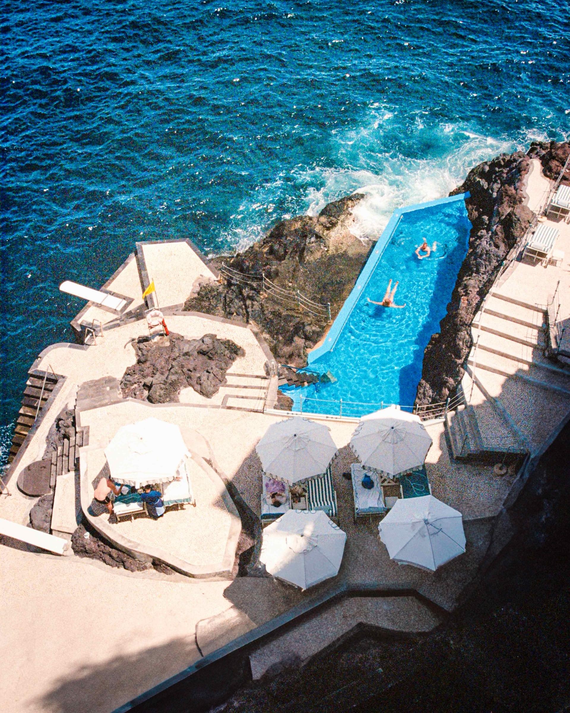 Seen from above, a narrow pool runs along a cliff edge into the sea, two people swimming in it, loungers and umbrellas on the terrace beside it.