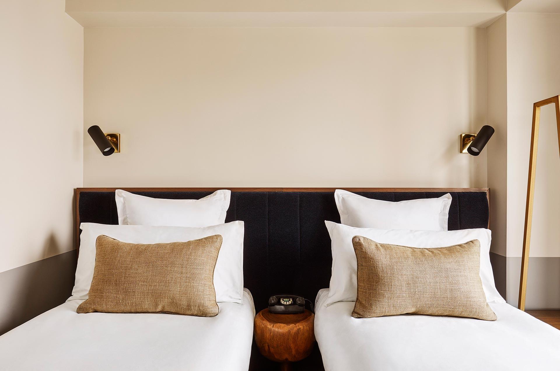 two twin beds with tan pillows against a dark upholstered headboard, with a black rotary phone on a round wooden table between them
