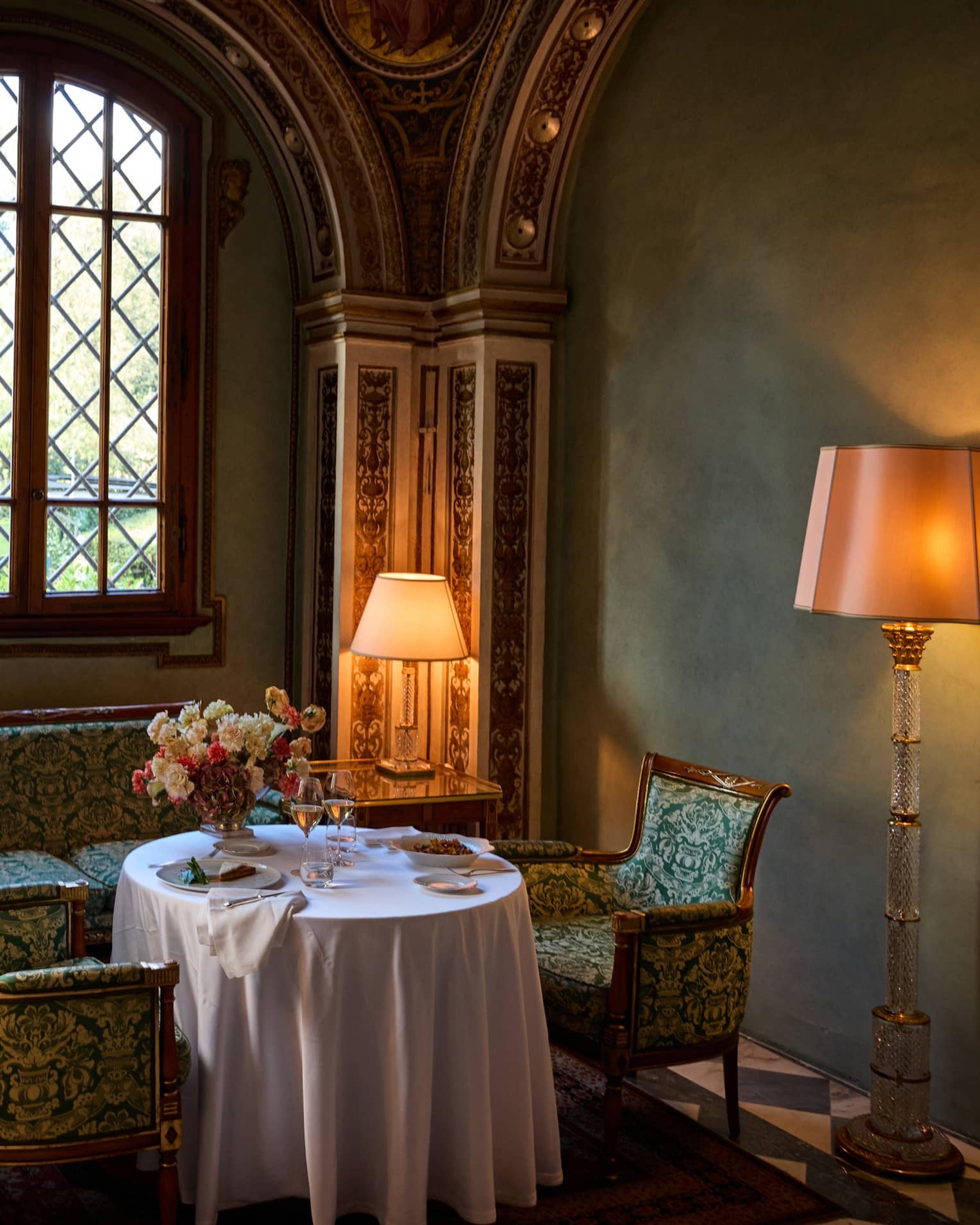 A round table set with plated food, wine glasses and flowers in a frescoed alcove, with a floor lamp and a window onto greenery.