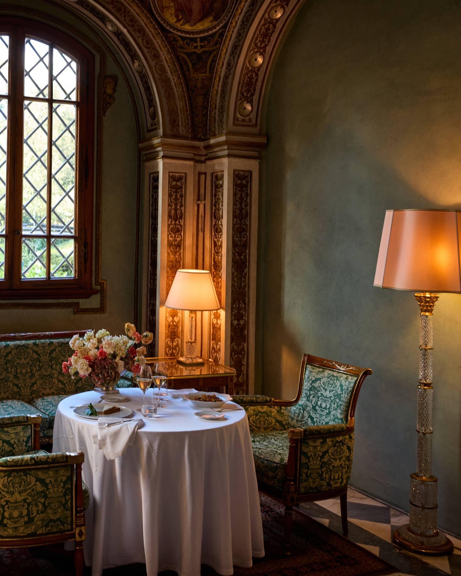 A round table set with plated food, wine glasses and flowers in a frescoed alcove, with a floor lamp and a window onto greenery.