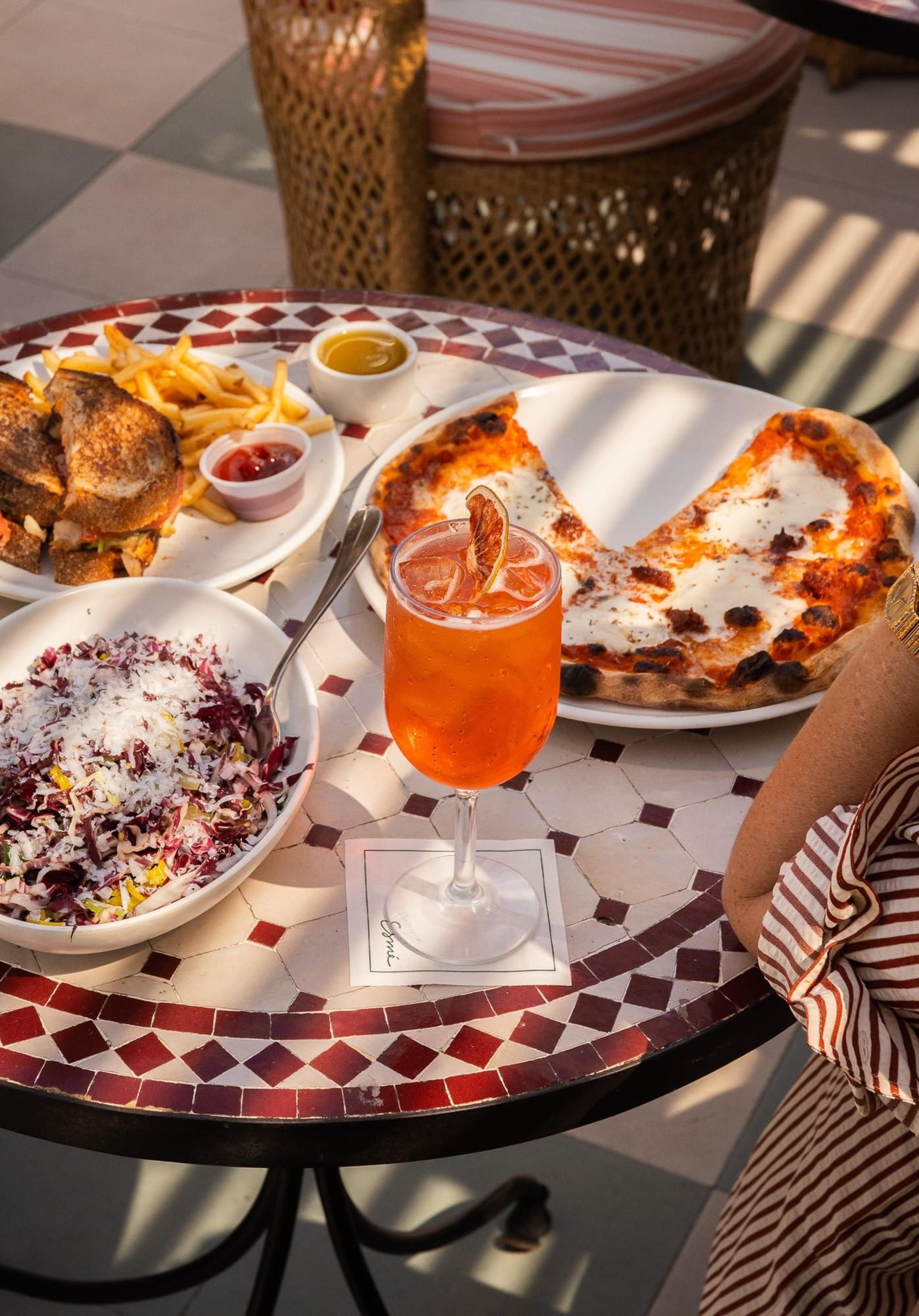 A round mosaic table set with a grilled sandwich and fries, a bowl of shaved salad, a wood-fired pizza, and an orange spritz in a stemmed glass, with a seated diner's arm at the edge.