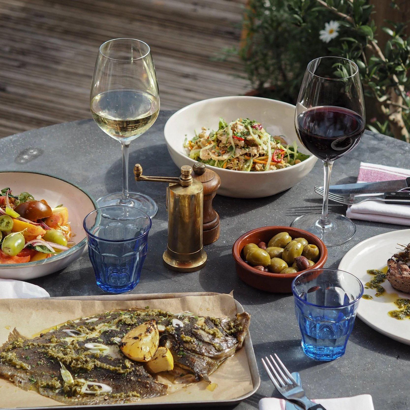 A table laid outdoors with glasses of white and red wine, bowls of salad and olives, and a tray of grilled whole fish with lemon.