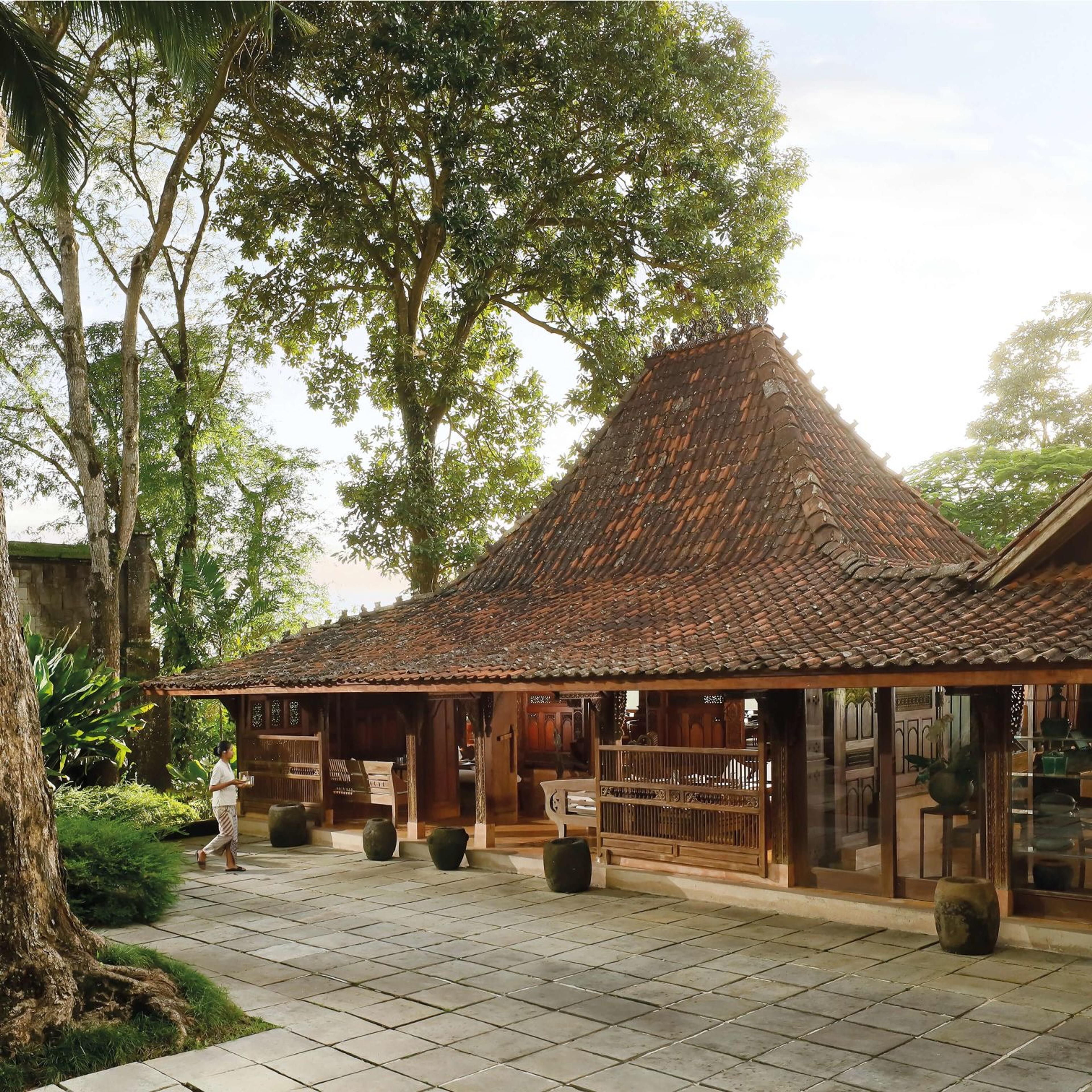 A staff member carrying a tray walks across a stone courtyard in front of a tiled, pitched-roof building under large trees.