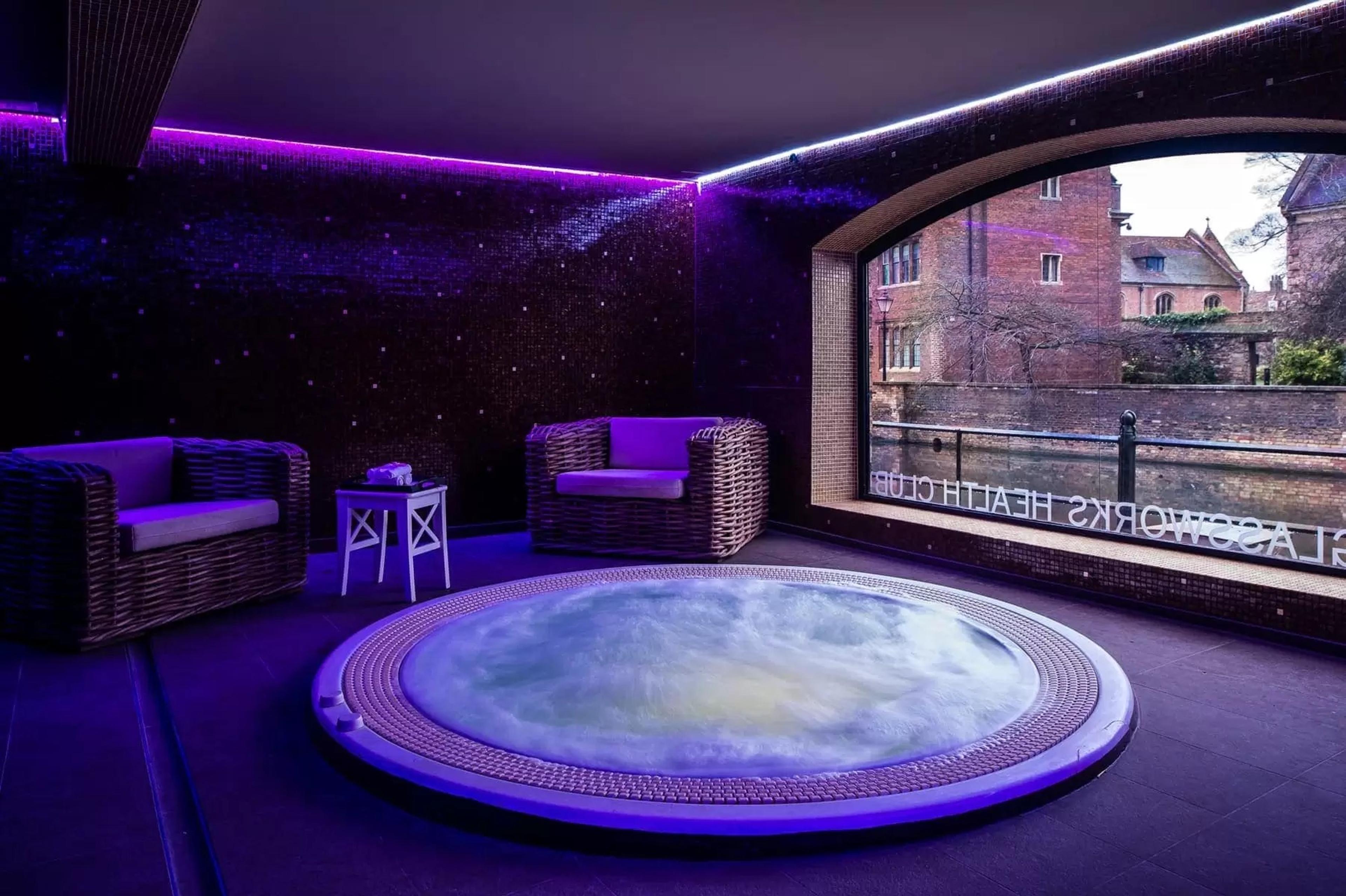 a round outdoor jacuzzi lit purple at night, with two wicker armchairs beside it and a view of a brick building through an arched window