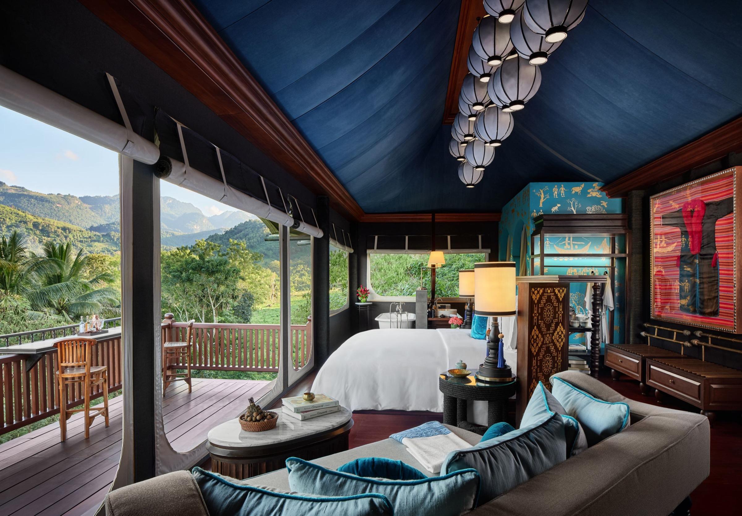A hotel suite with a blue draped ceiling and paper lantern lights, a white bed, a sofa with teal cushions, and an open-sided deck looking over forested hills.