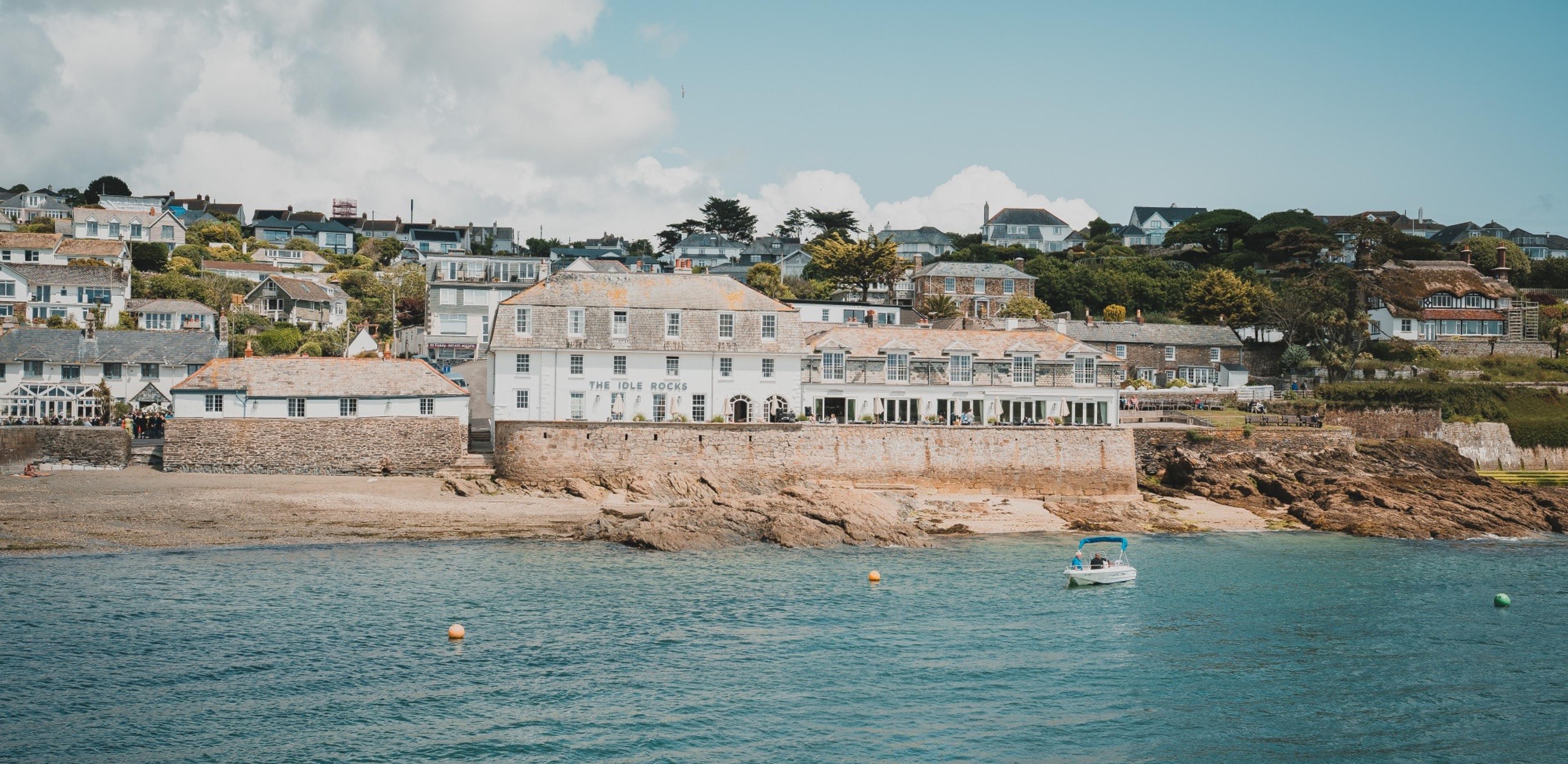 The same white harbourside hotel seen from the water in daylight, with a small boat moored offshore, a beach exposed at low tide, and houses on the hillside behind.