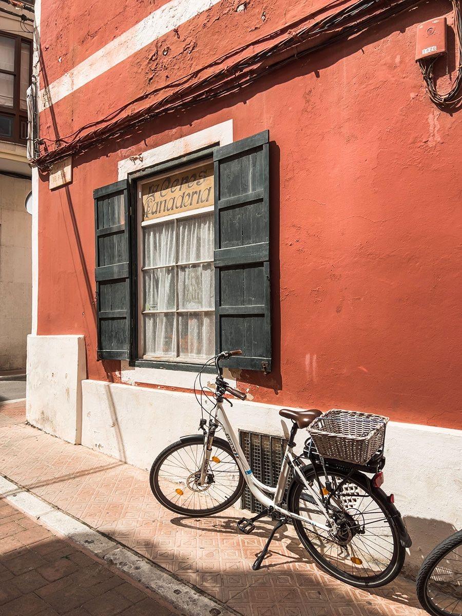 A white bicycle with a wicker basket parked against a red-painted building with a shuttered window reading Panaderia.