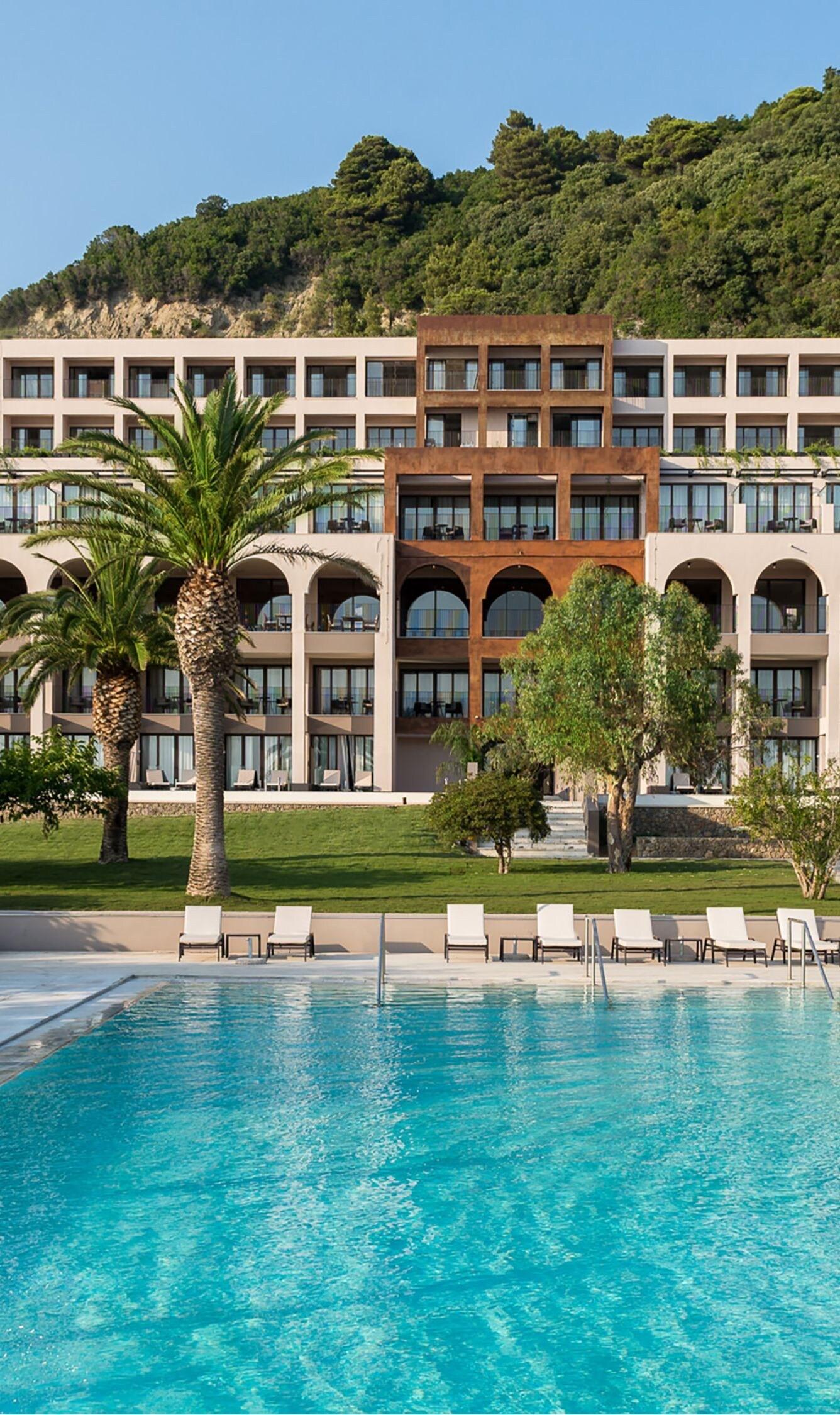 A multi-storey cream hotel building with a rust-colored accent facade, backed by a pine-covered hillside, with a turquoise pool and palm trees in the foreground.