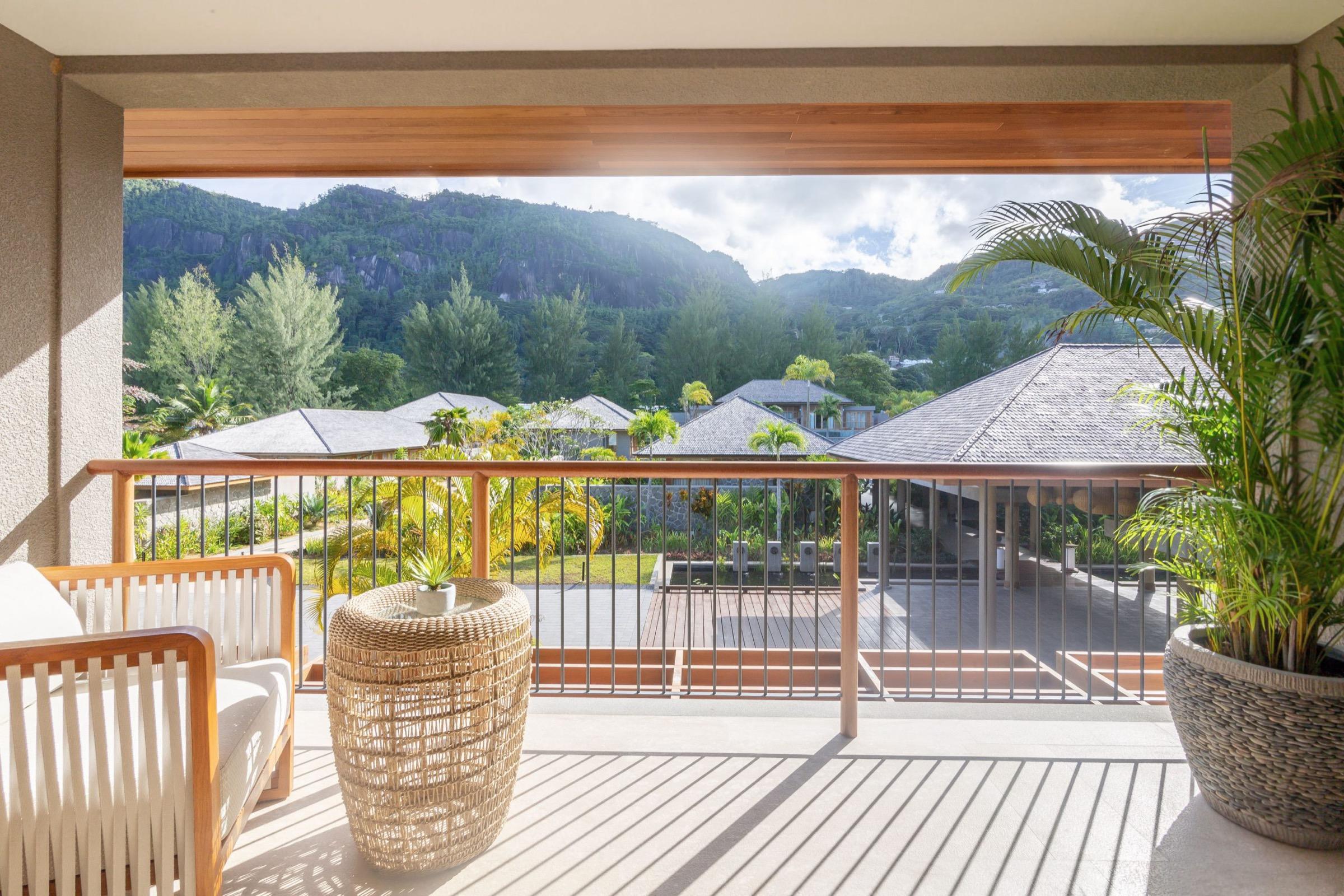A balcony with a rattan sofa and stool, looking out over resort rooftops to forested hills.