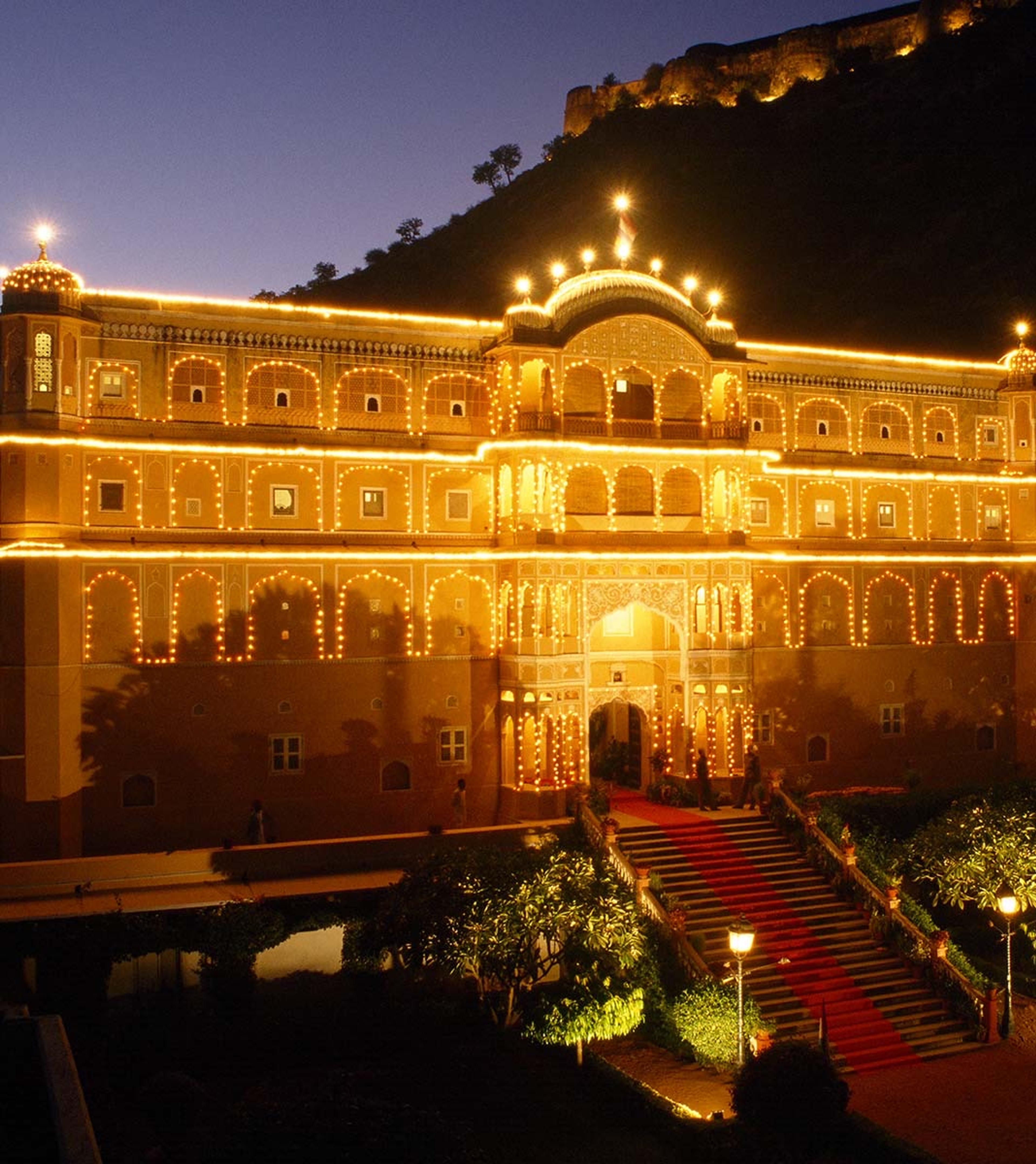 The palace's arched facade lit up at night against a hillside, with a red carpet staircase leading up to the entrance.