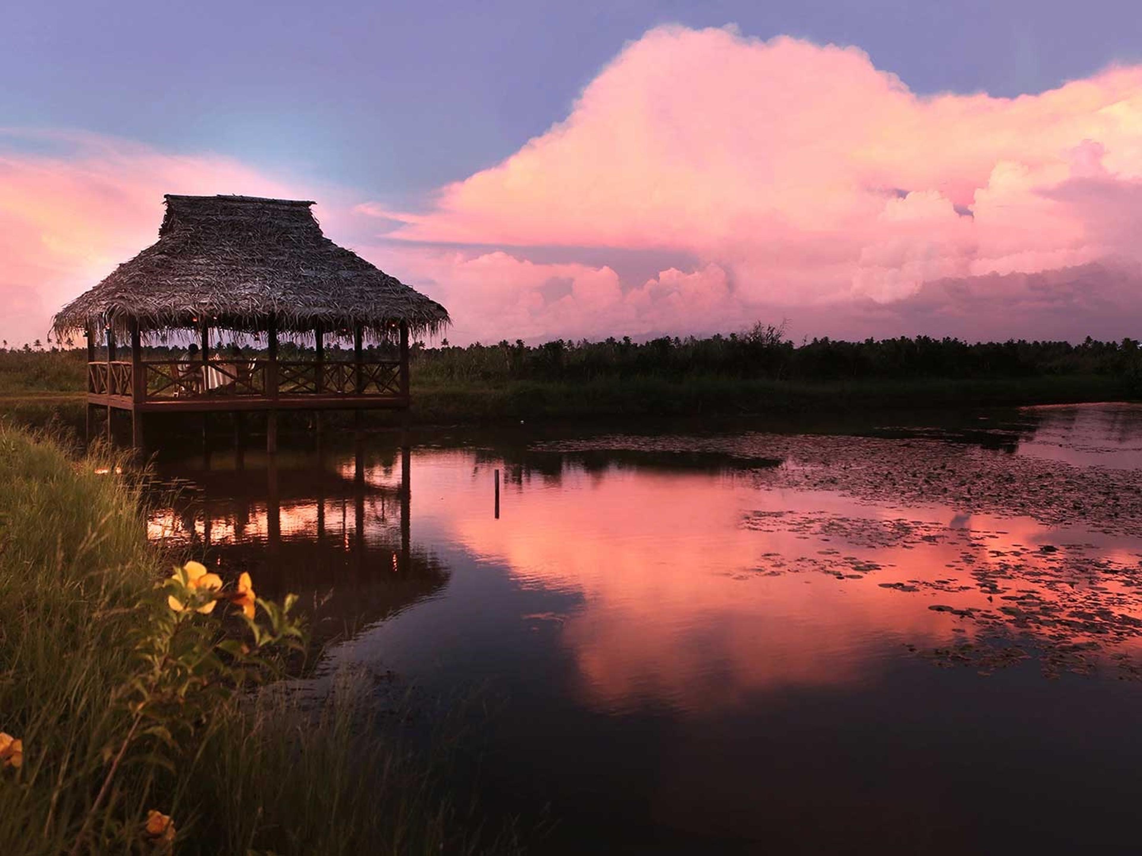A thatched pavilion over a still lagoon at sunset, the sky pink in the water