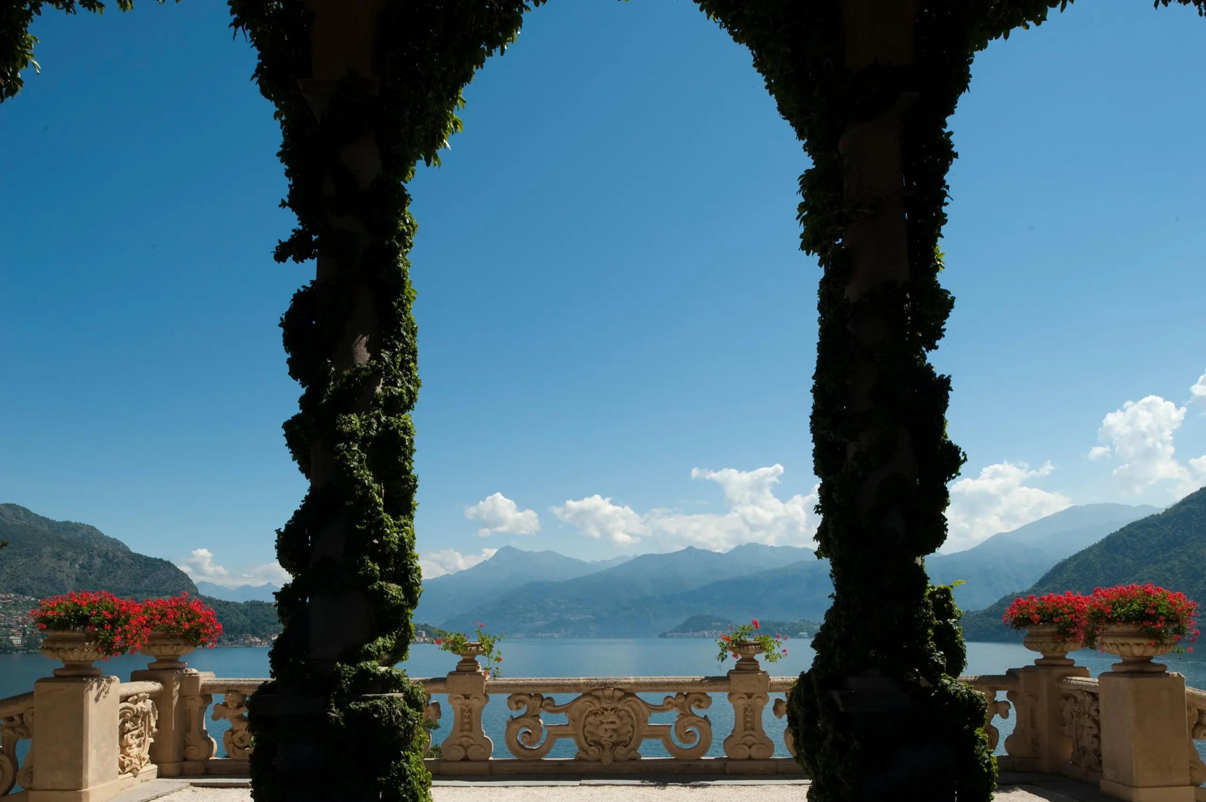 Stone columns wrapped in ivy frame a view of Lake Como and the mountains beyond a balustrade lined with potted red flowers.