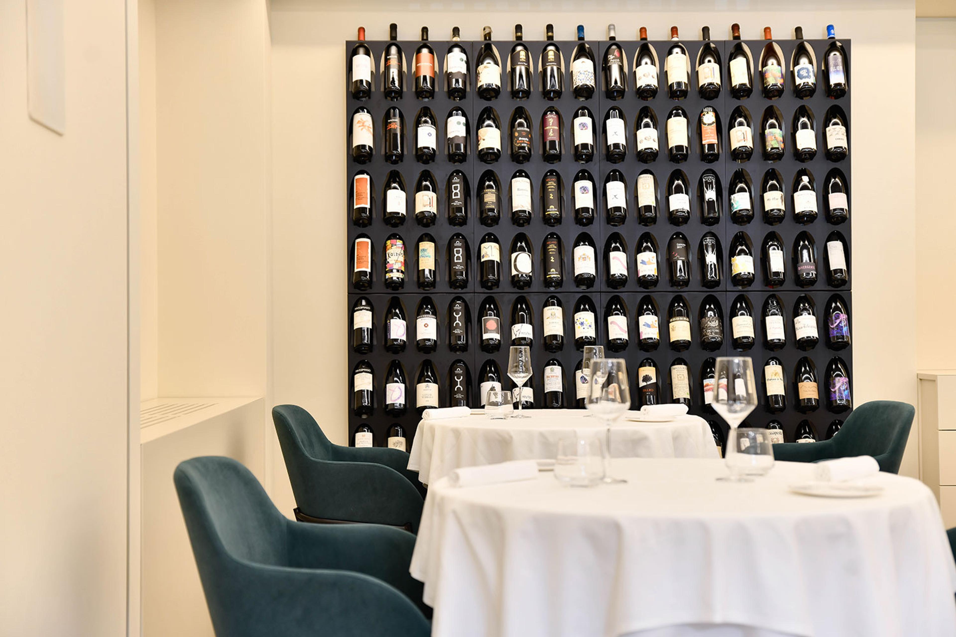 White-clothed tables and green velvet chairs in a small dining room, a wall of wine bottles behind