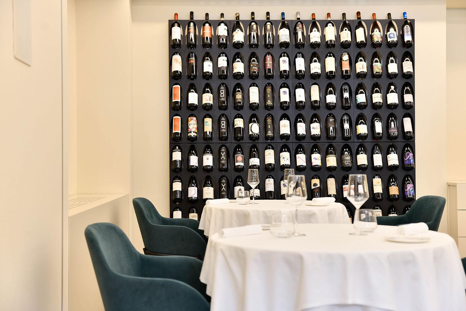 White-clothed tables and green velvet chairs in a small dining room, a wall of wine bottles behind