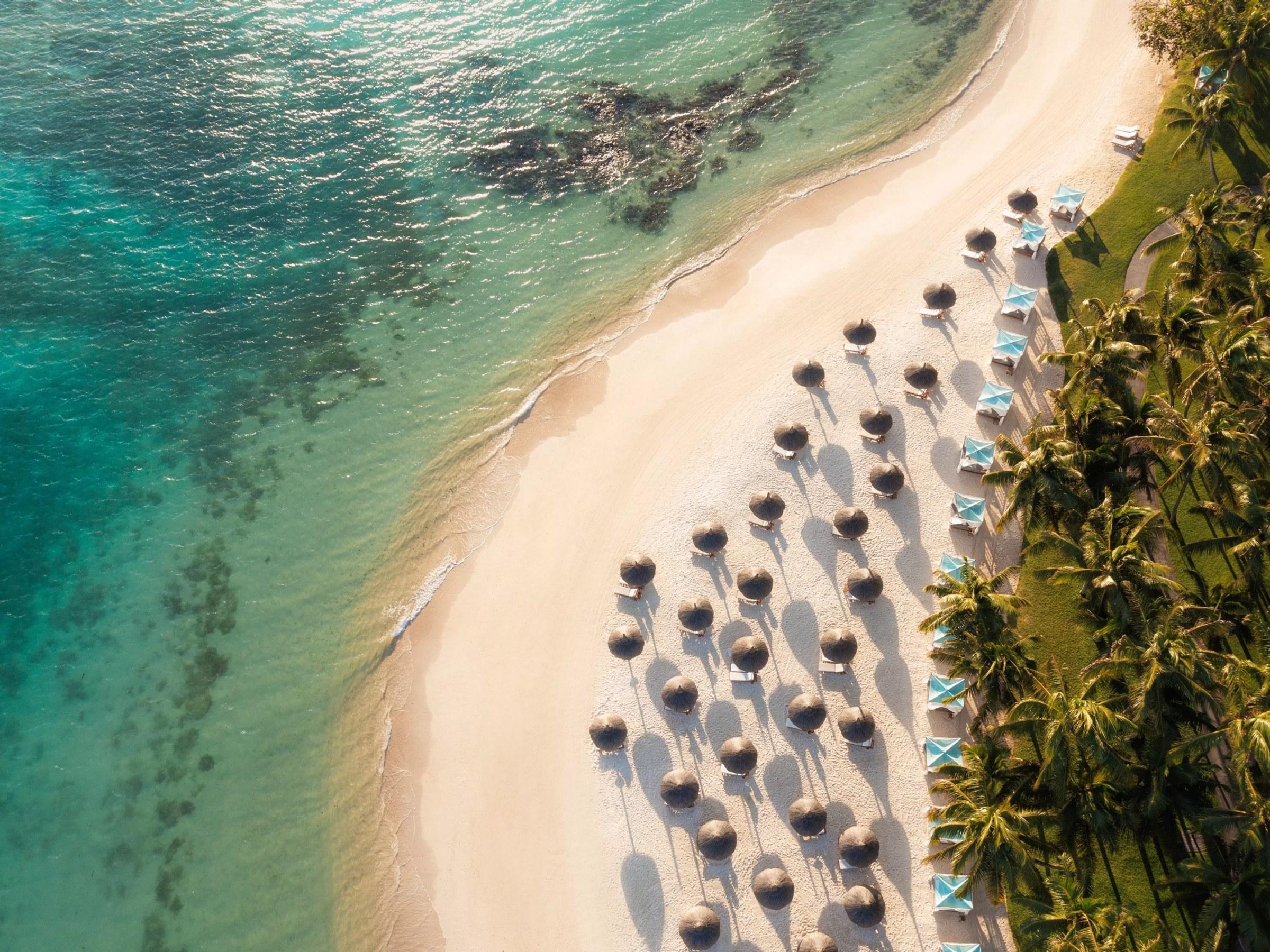 A curve of white sand and turquoise lagoon seen from above, lined with thatched parasols and palms