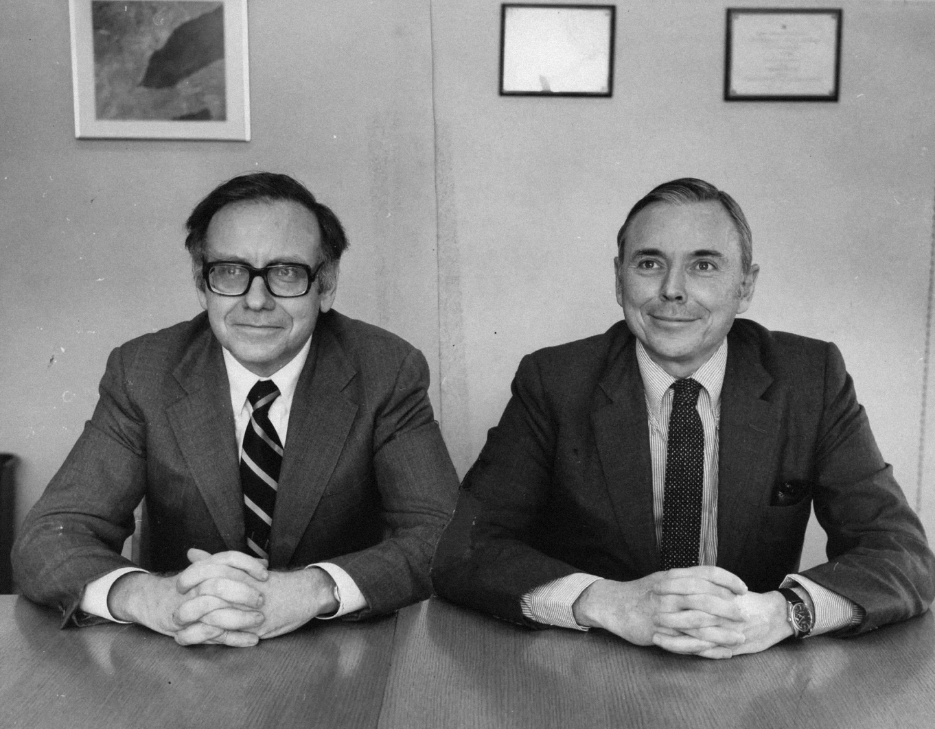 A black and white photograph of two men in business suits seated at a table, taken likely in the 1970s. On the left is a man wearing thick-rimmed glasses and a striped tie. On the right is a man wearing a polka dot tie and striped shirt. Both men are smiling slightly and have their hands clasped on the table. There are framed items hanging on the wall behind them.