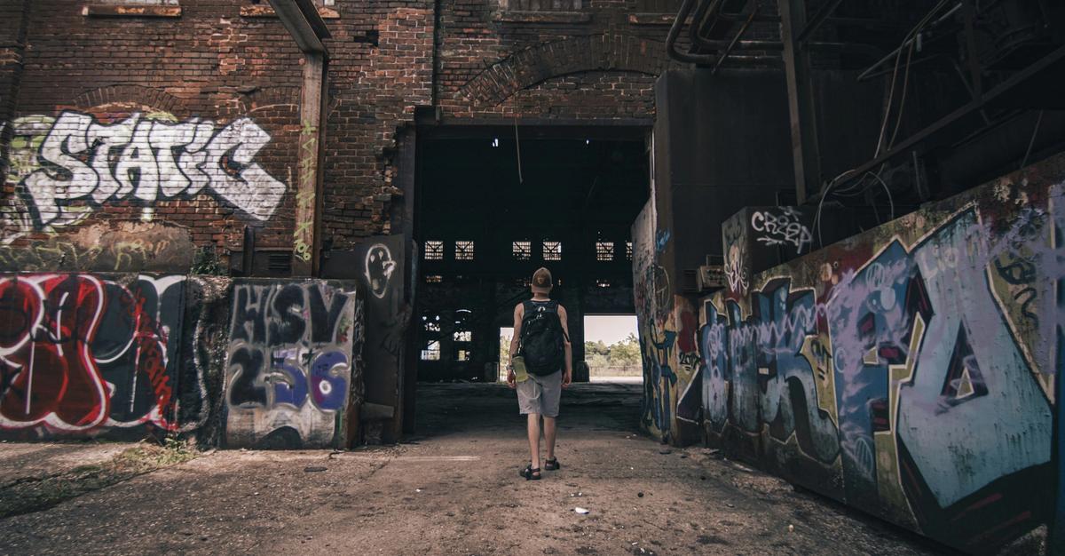 Man with back pack walking into an abandoned factor with graffiti.